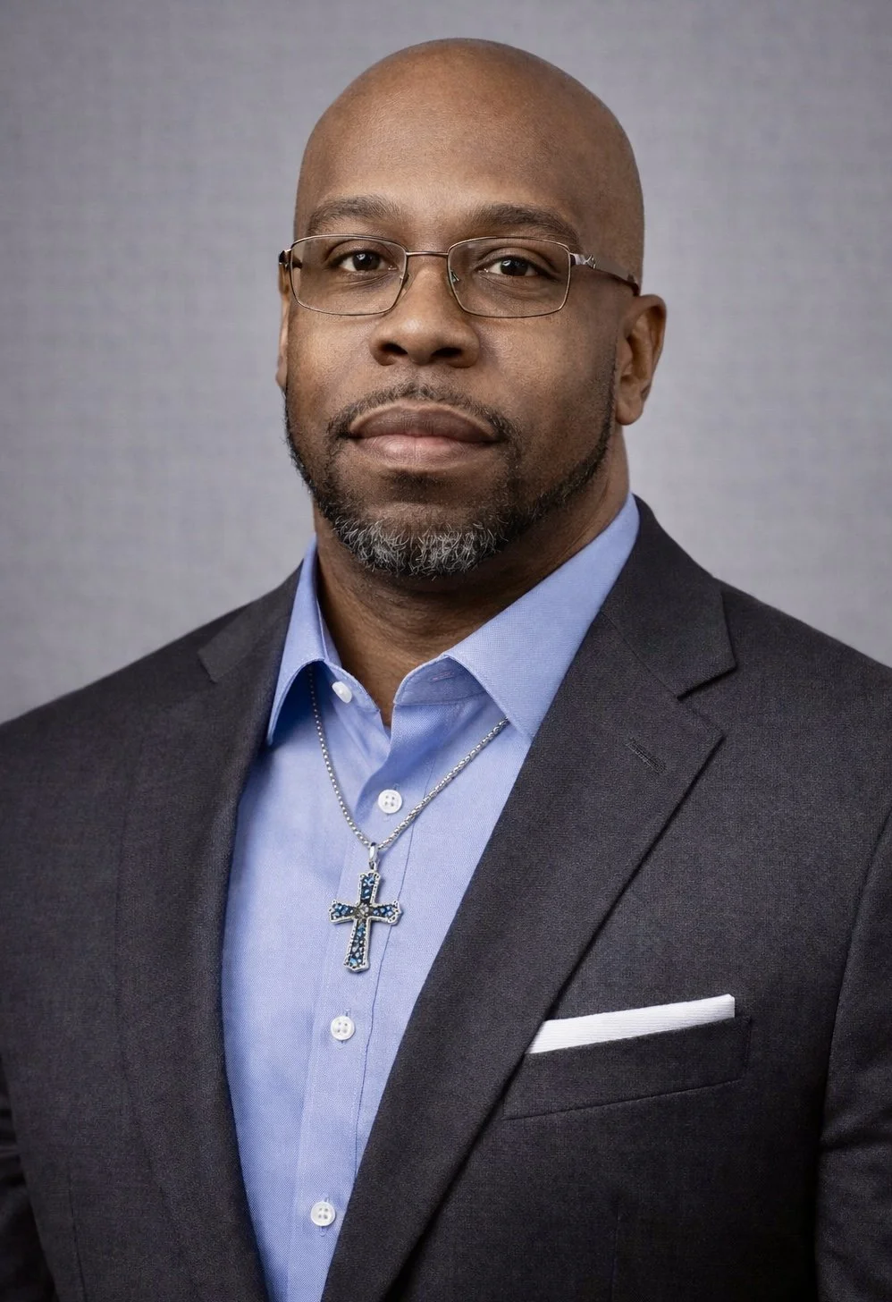 A man with glasses, a beard, and a shaved head wearing a dark suit, light blue shirt, a cross necklace, and a white pocket square in front of a gray background.