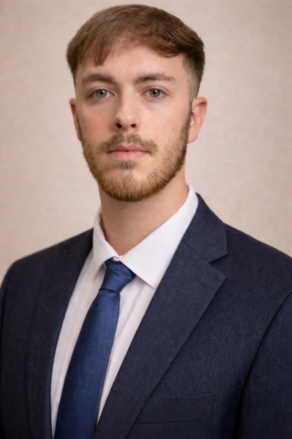 A young man with light skin, short brown hair, and green eyes wearing a dark suit, white shirt, and blue tie, looking directly at the camera against a plain beige background.