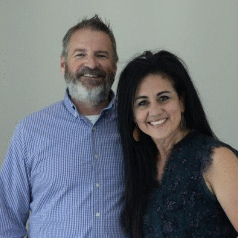 A man and woman smiling and standing close together, with a plain wall in the background.