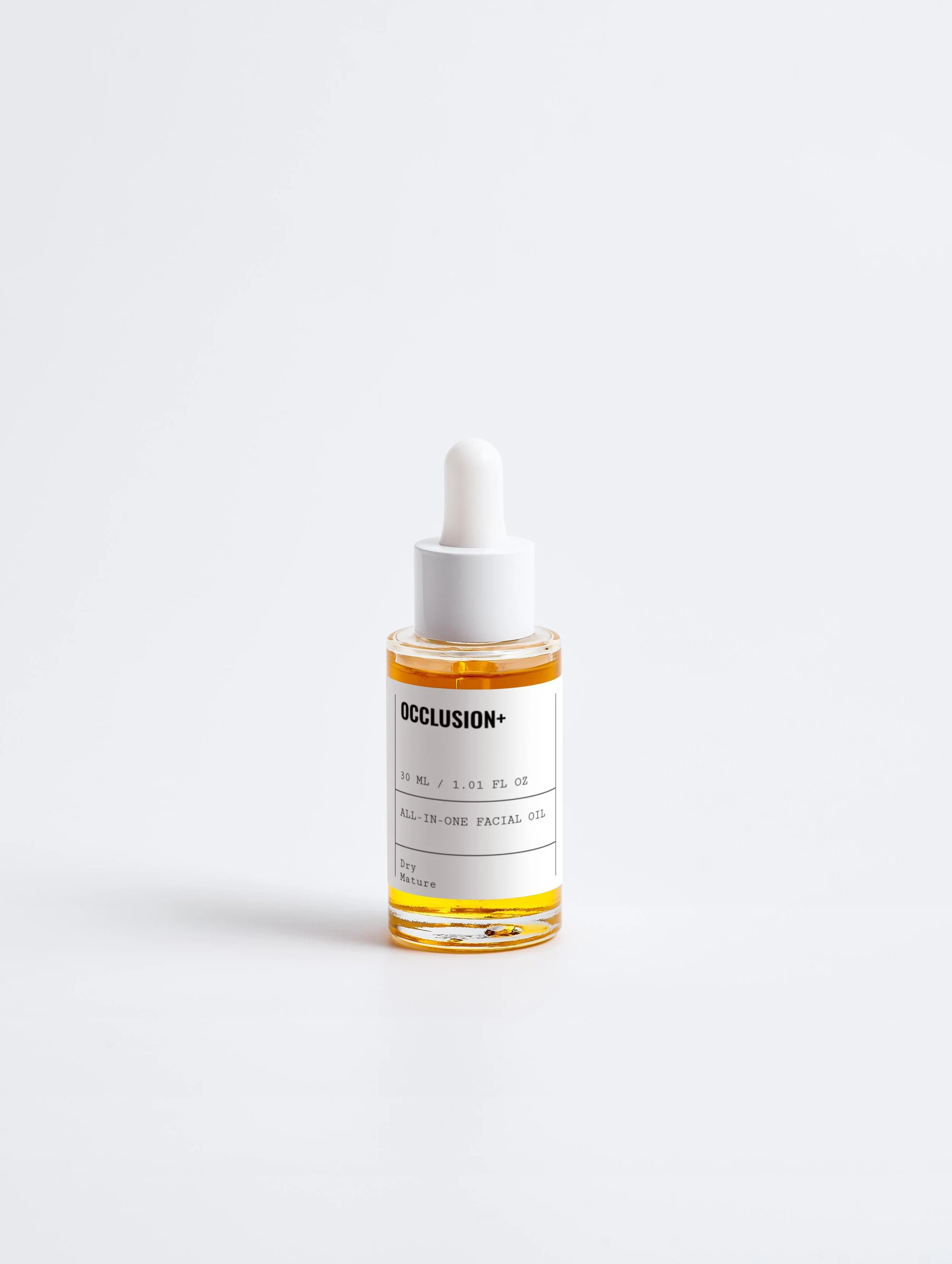 A small glass bottle with a white dropper cap, containing golden facial oil labeled "OCCLUSION+" and "All-in-One Facial Oil" on a plain white background.