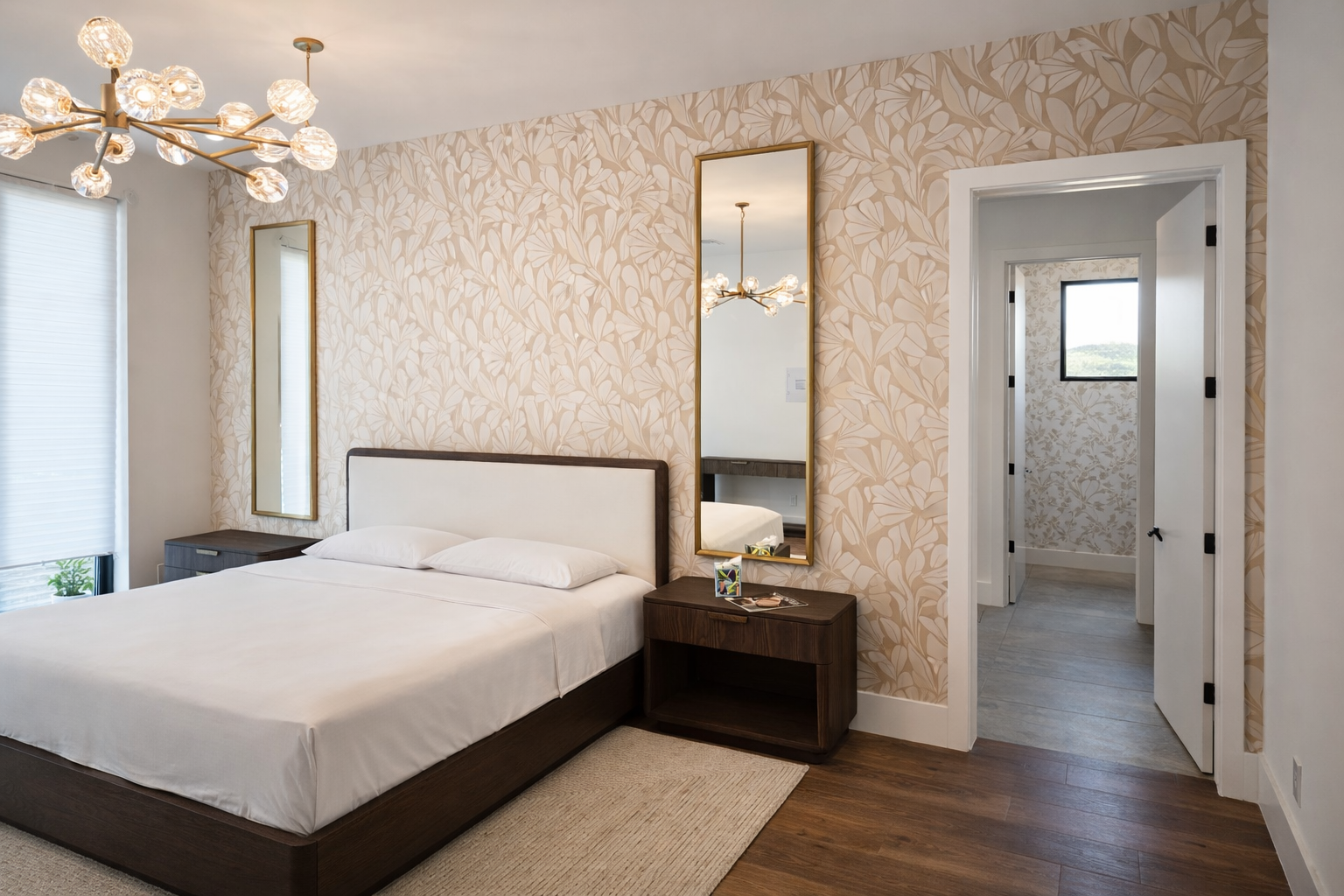 Modern bedroom with floral wallpaper, a bed with white bedding, and a wooden nightstand. There are two tall mirrors on the wall, a stylish chandelier, and a window with blinds.