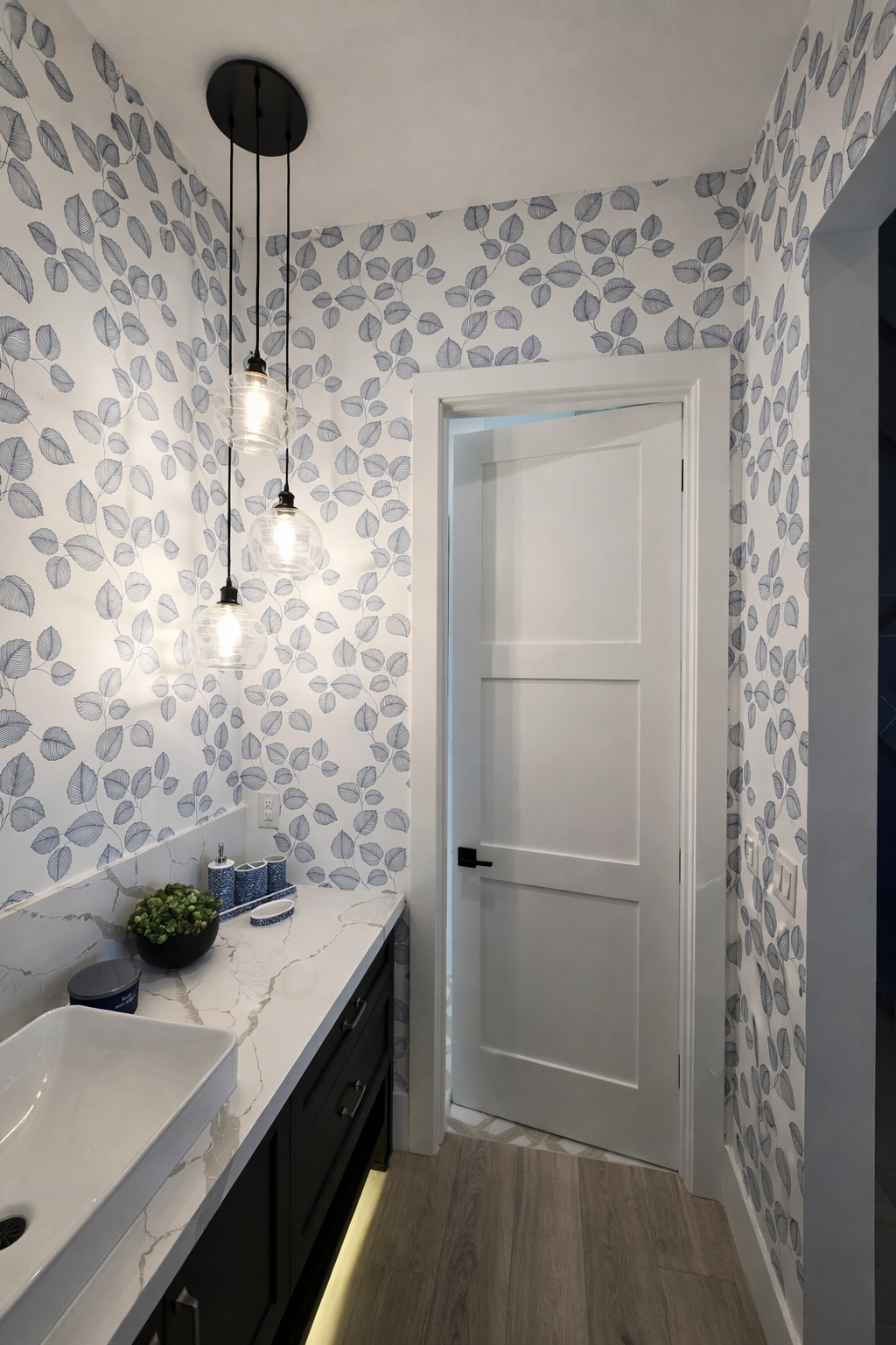 A modern laundry room with leaf-patterned wallpaper, a white door, black cabinetry, and a marble countertop with a sink, decorated with a bowl of greenery and blue containers. Three hanging pendant lights illuminate the space.