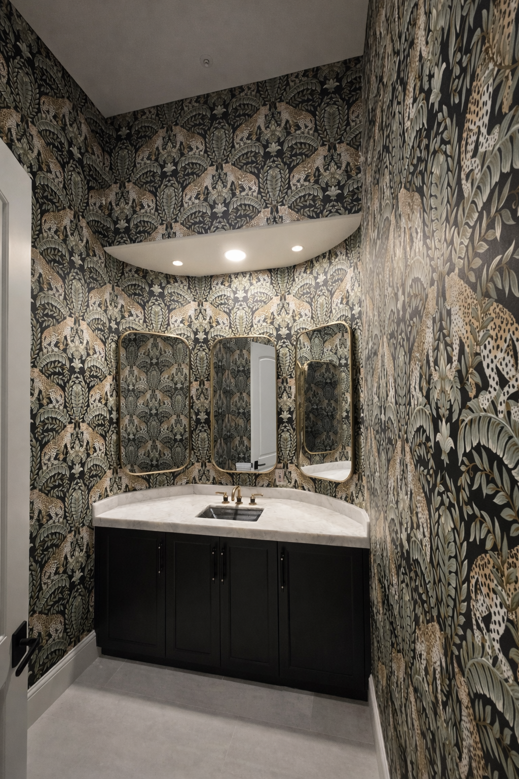 Small bathroom with patterned wallpaper featuring leopards and mythical animals, black vanity with a white marble countertop, three mirrors with gold frames, and a small sink with gold fixtures.