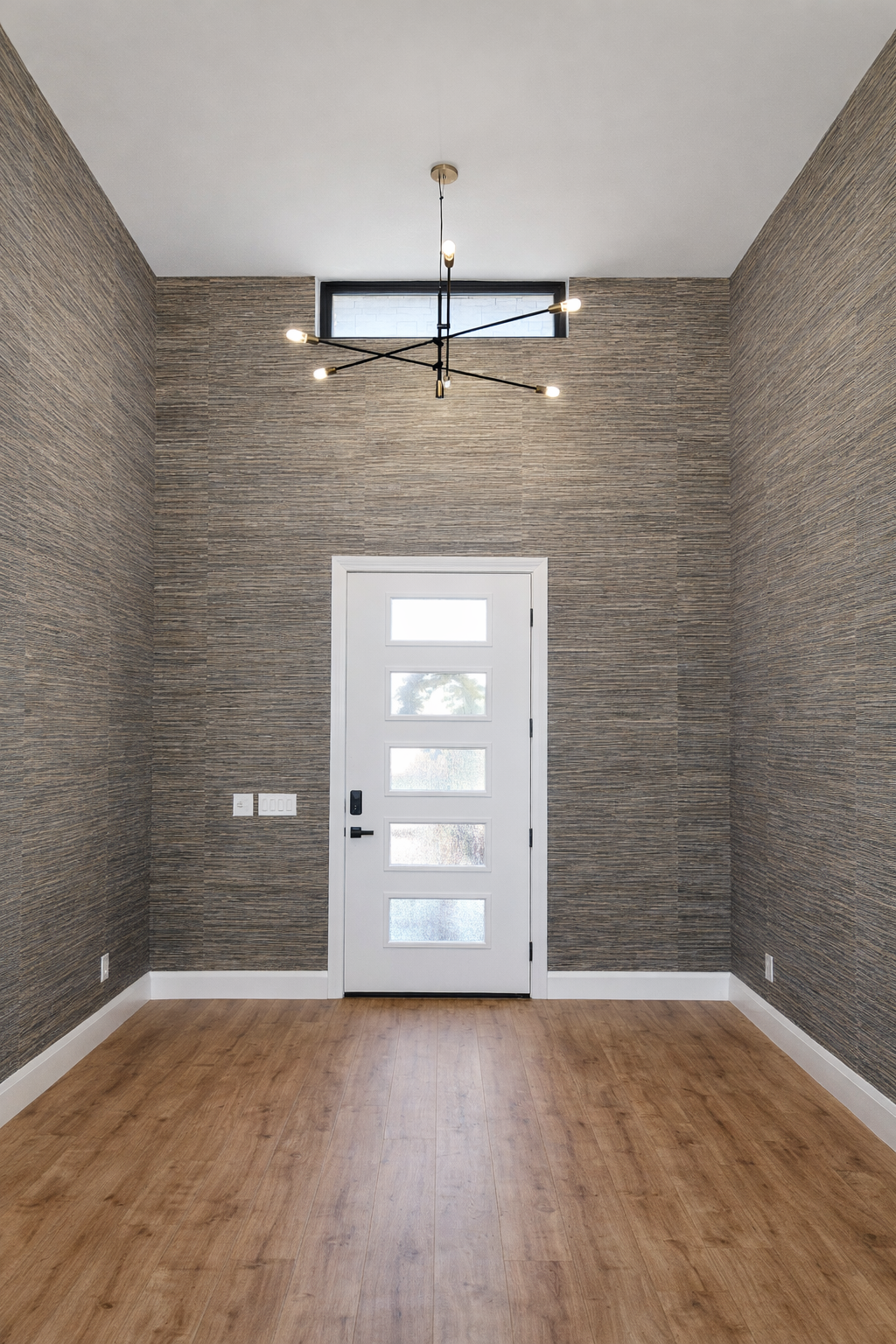 Empty interior room with a white front door, textured brown walls, wood flooring, a modern black chandelier, and a large window in the ceiling.
