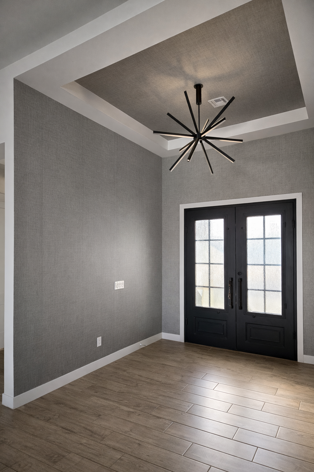 Empty modern room with gray textured walls, wood flooring, black double doors with glass panes, and a black geometric chandelier hanging from the ceiling.
