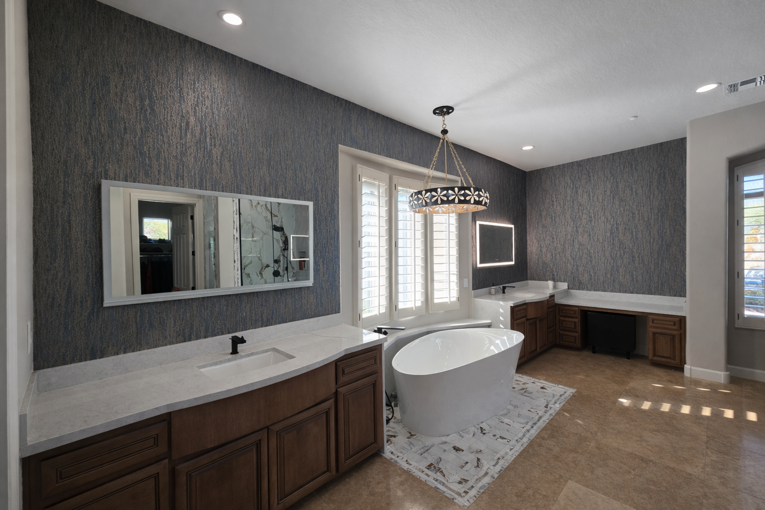 Modern bathroom with a freestanding bathtub, vanity with wooden cabinets, a large mirror, and a contemporary chandelier. The room features dark textured walls, white plantation shutters, and a tiled floor.