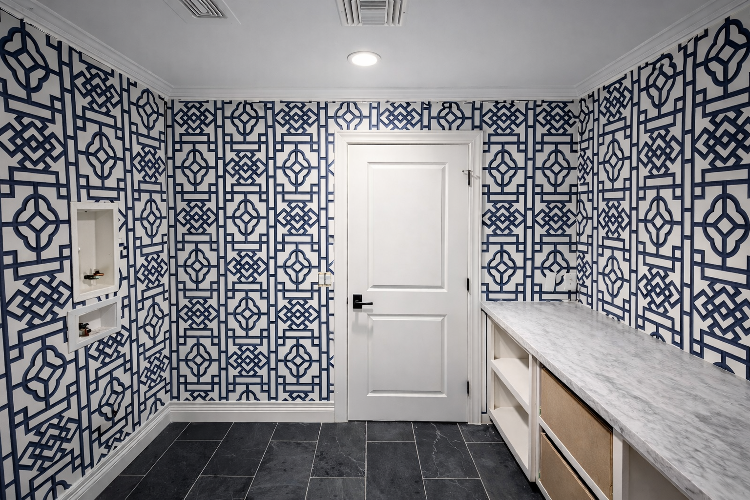 Interior of a room with patterned blue and white wallpaper, black tiled floor, a white door, and a marble countertop with open shelves underneath.