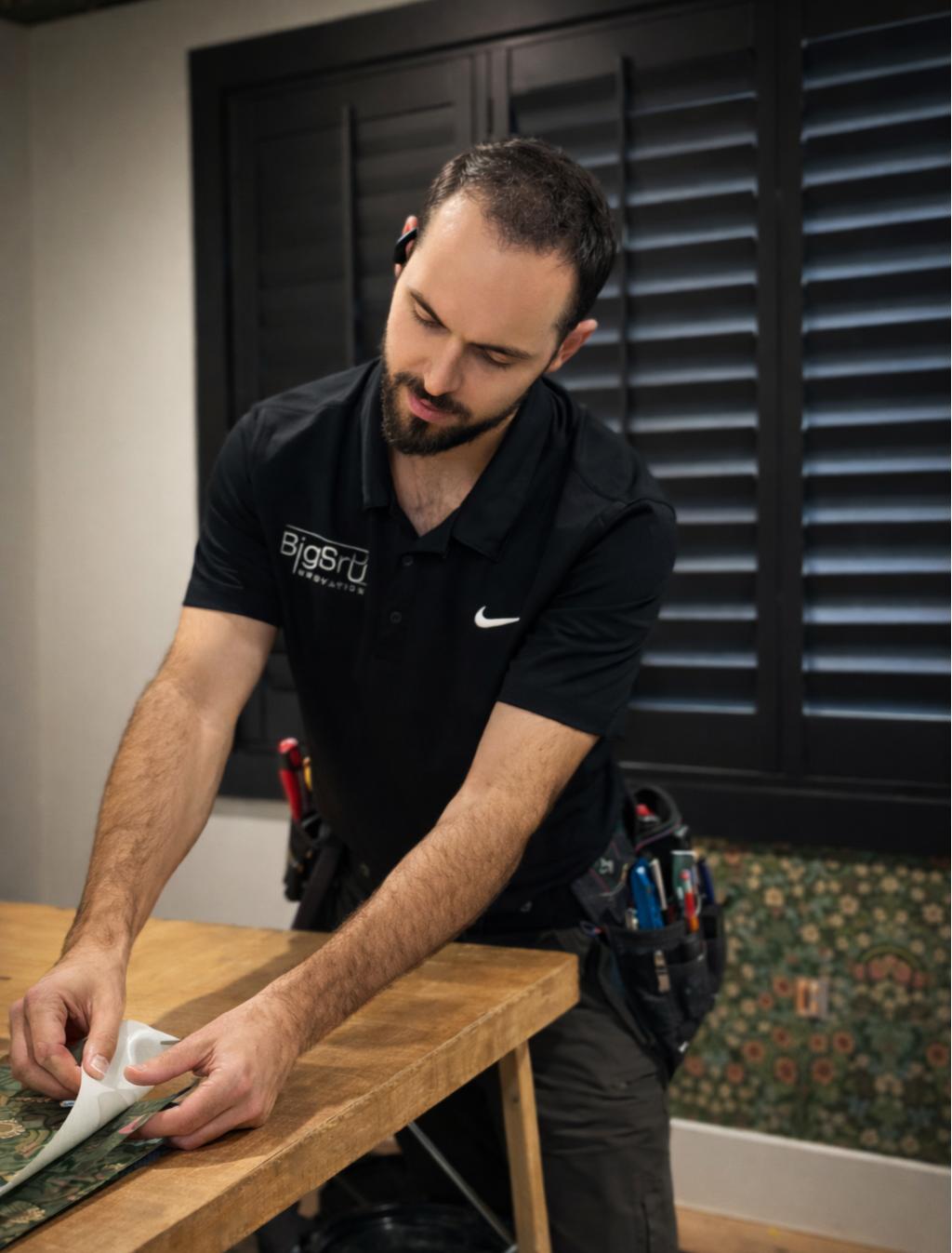 A man with a beard, wearing a black polo shirt with company logo, is working on a wooden table, prepping wallpaper in a room with dark shutters.