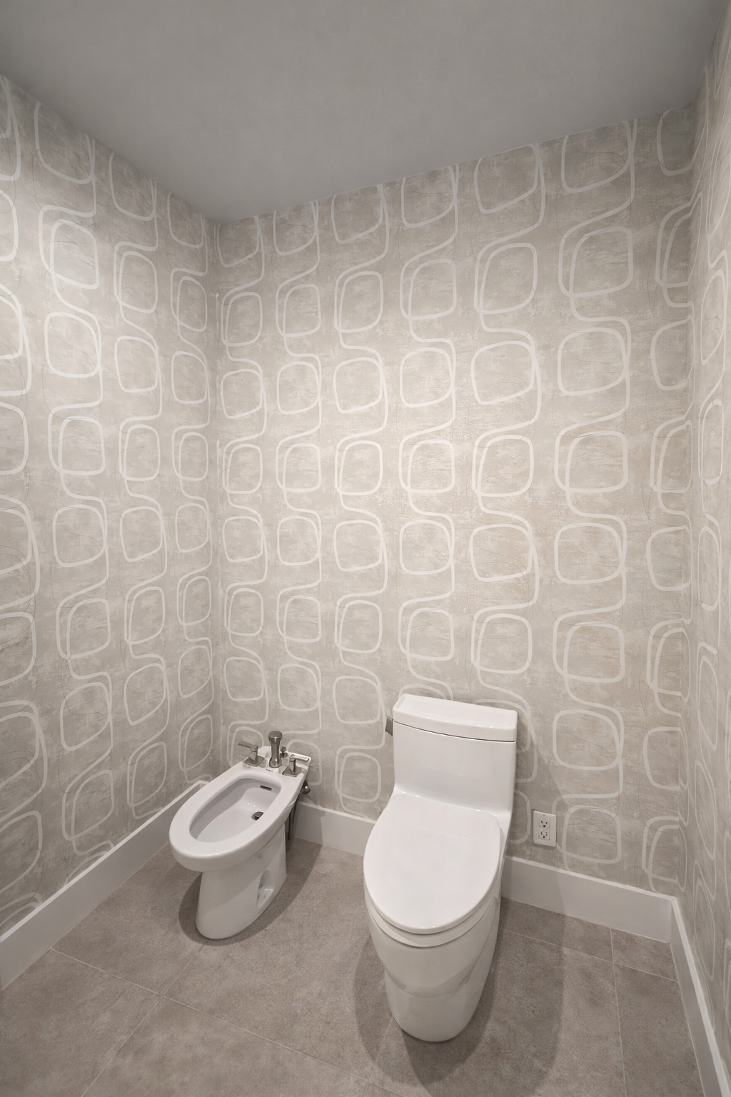 A small bathroom with a modern toilet and a bidet, surrounded by light colored walls with geometric line patterns and a gray tiled floor.