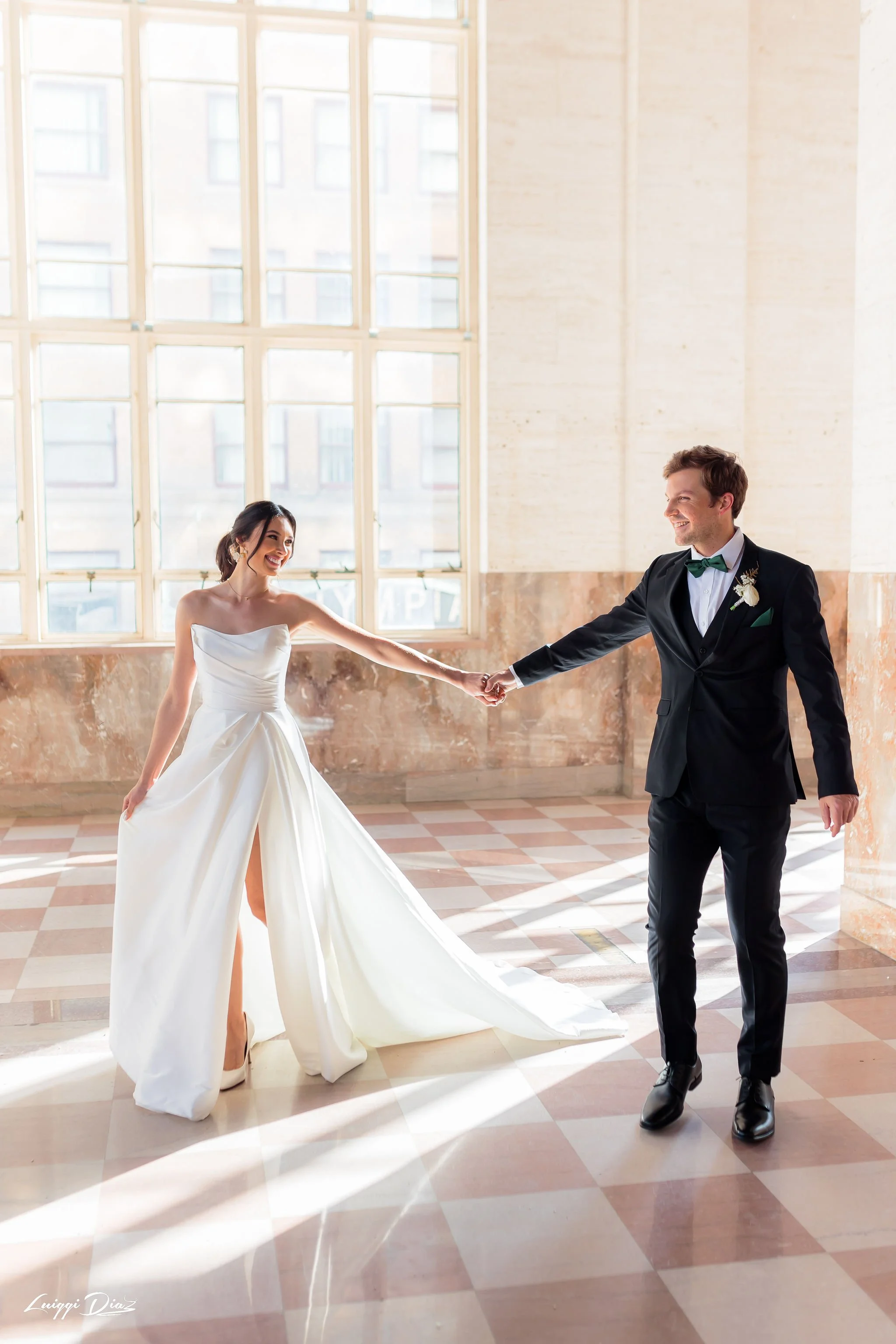 A bride and groom holding hands and dancing in a bright, spacious room with large windows and checkered tile floor.