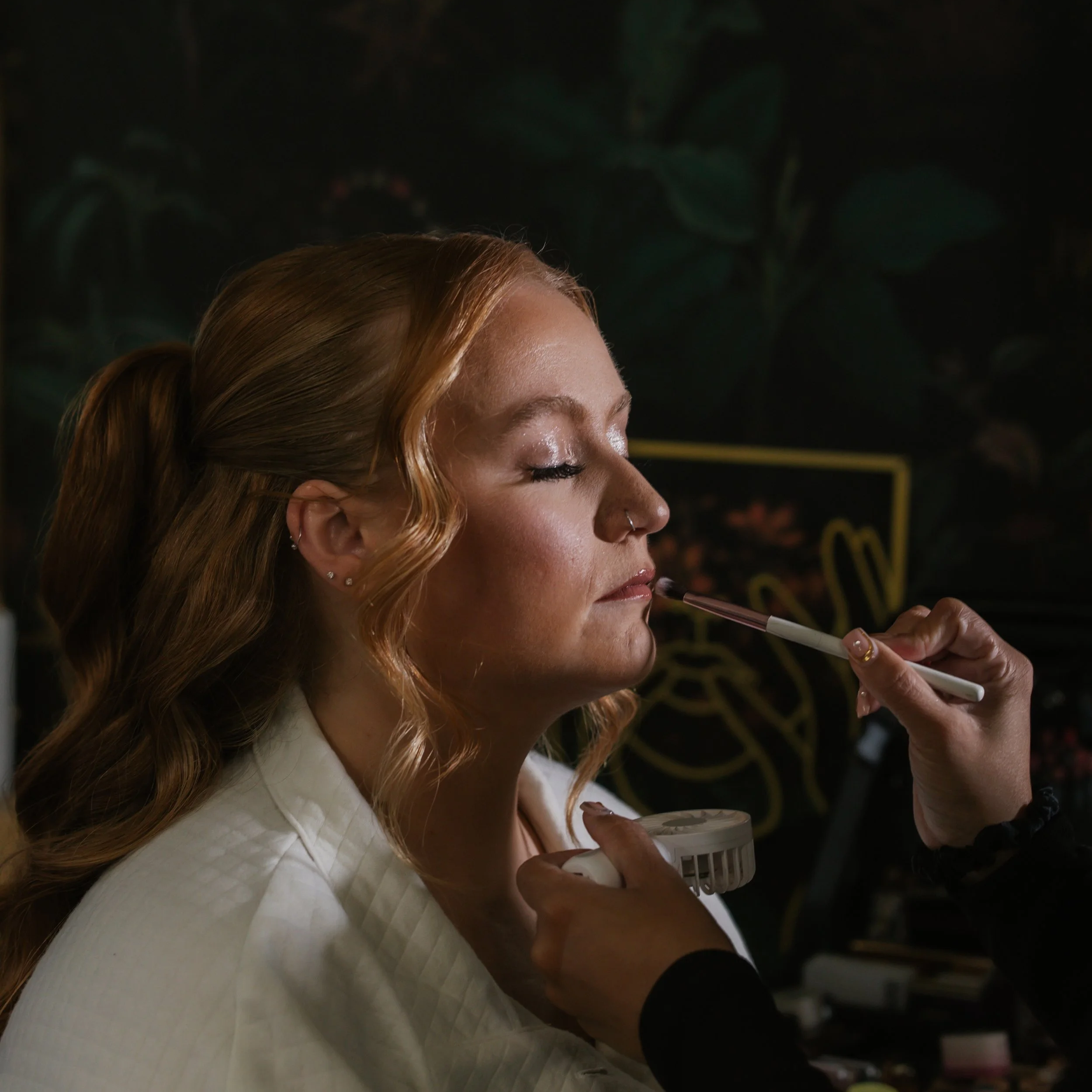 A woman with red hair getting her makeup done by a florida makeup artist in a dimly lit room.
