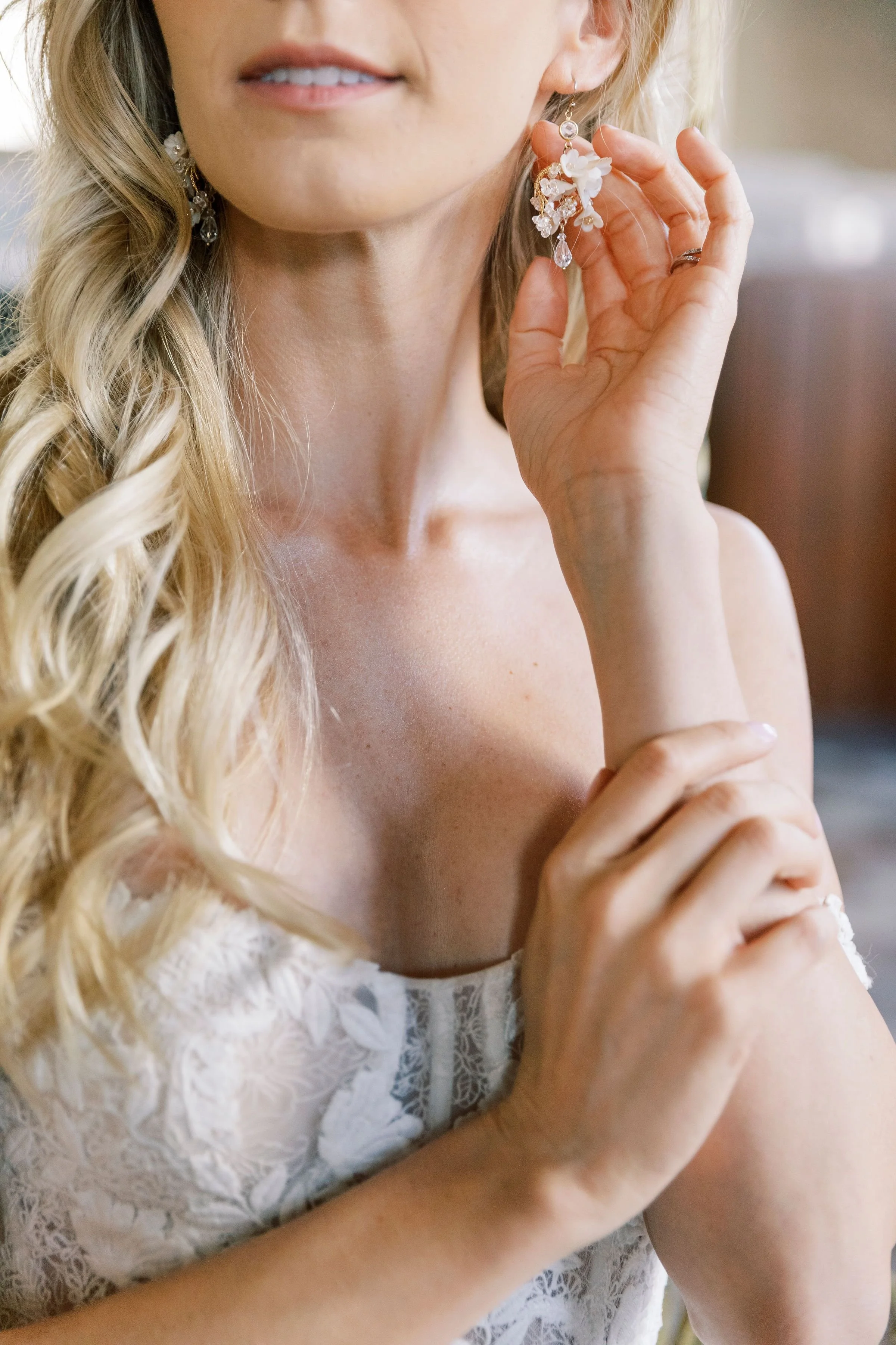 A woman is holding an ornate earring with floral and crystal details close to her ear, wearing a lace dress.