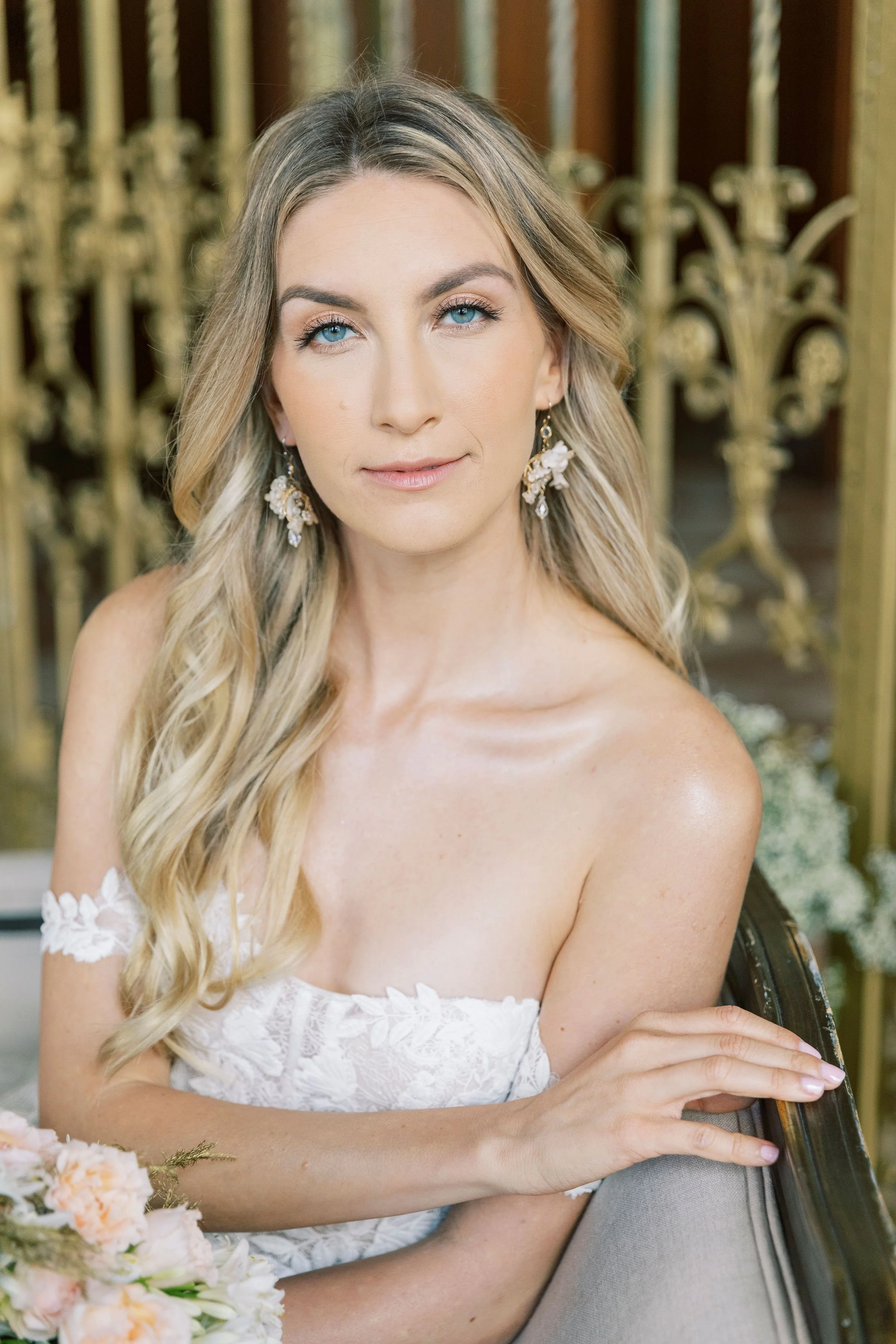 A woman with soft wedding makeup and wavy blonde hair wearing a white dress with lace details, floral earrings, and holding a bouquet of light-colored flowers, seated on a vintage chair in front of an ornate gold railing.