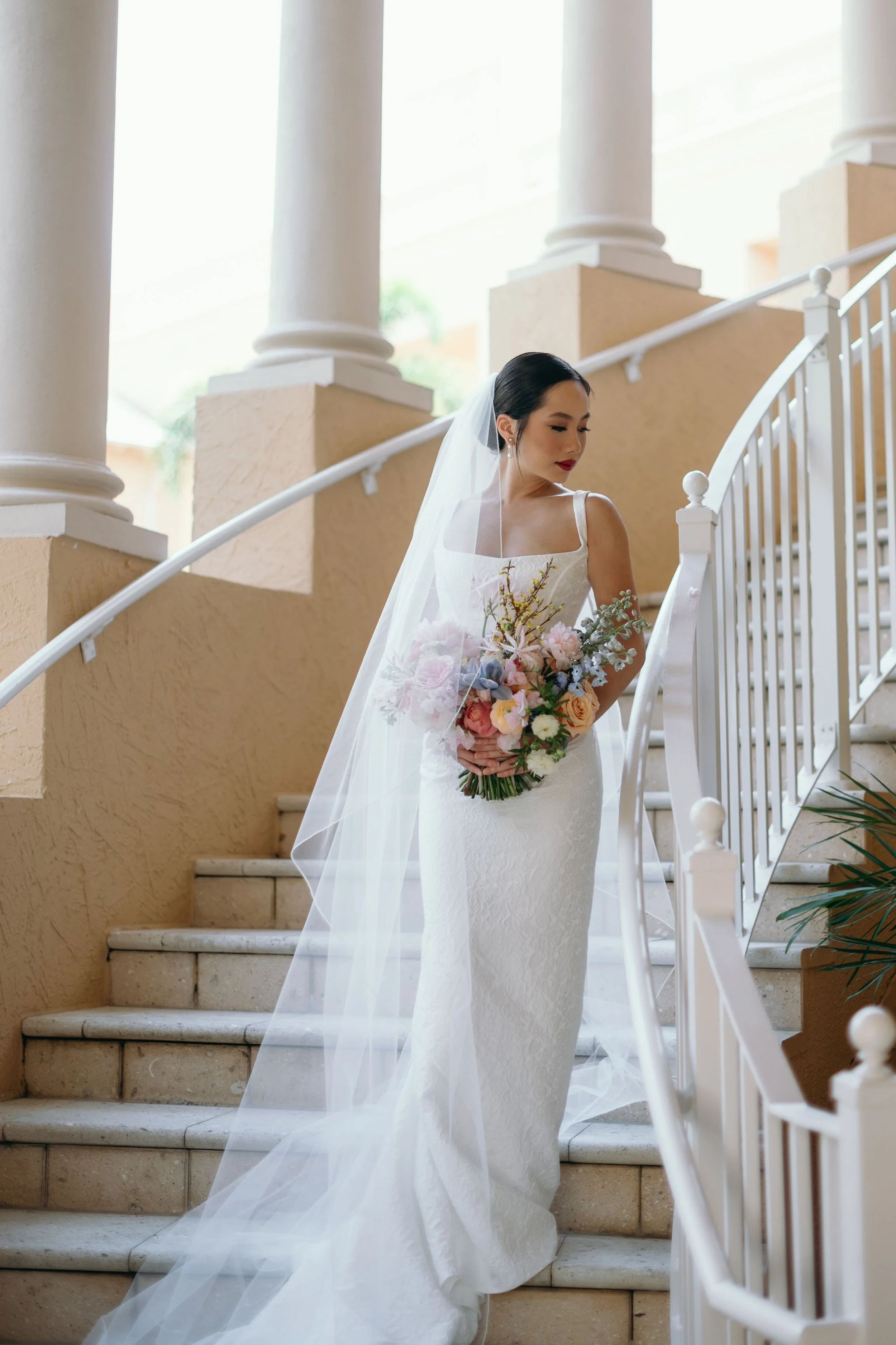 A bride standing on stairs indoors, wearing a white lace wedding gown with thin straps, holding a bouquet of pink, peach, and white flowers with greenery, and wearing a veil.