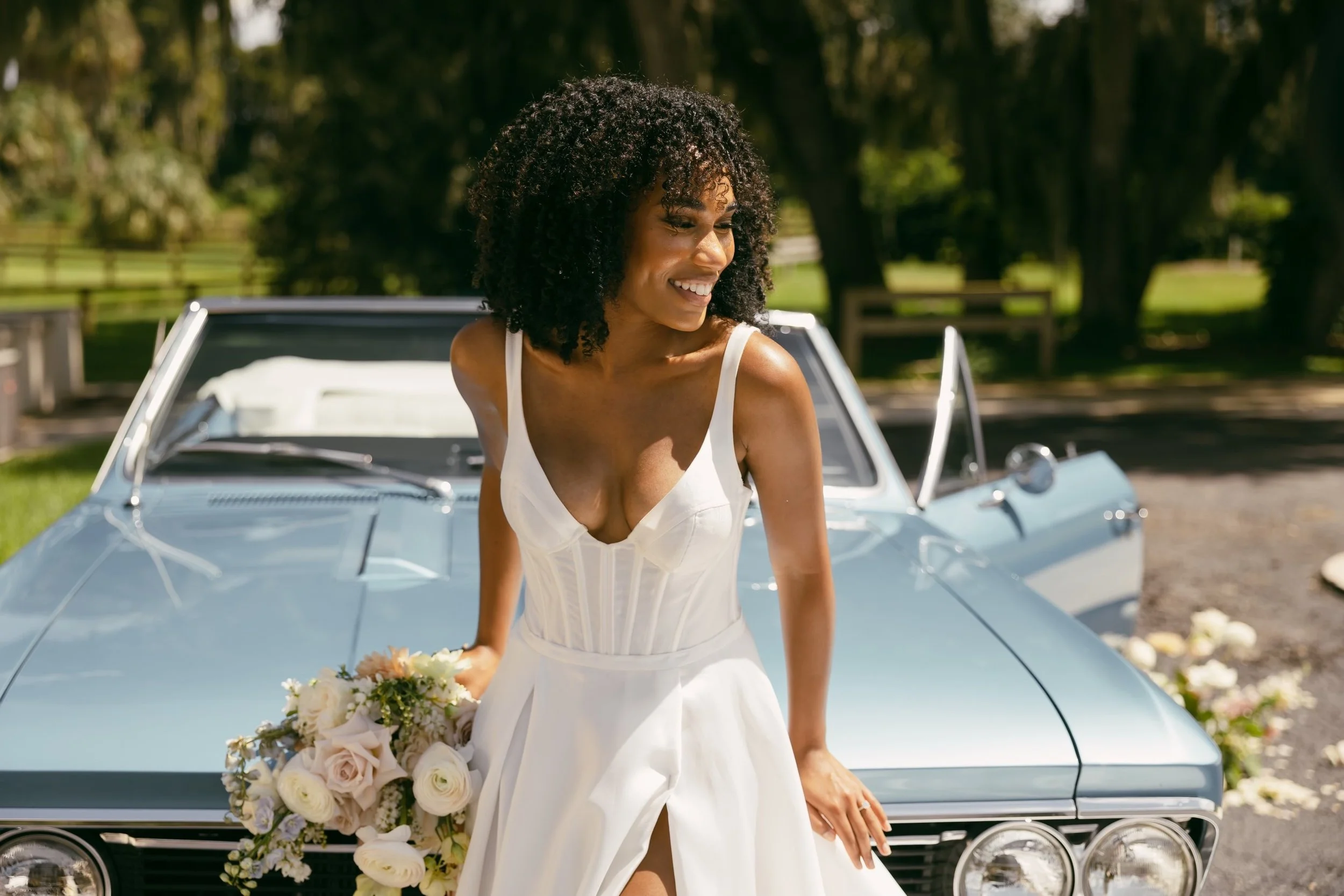 A woman in a white wedding dress holding a bouquet of flowers, standing in front of a vintage blue car outdoors on a sunny day.