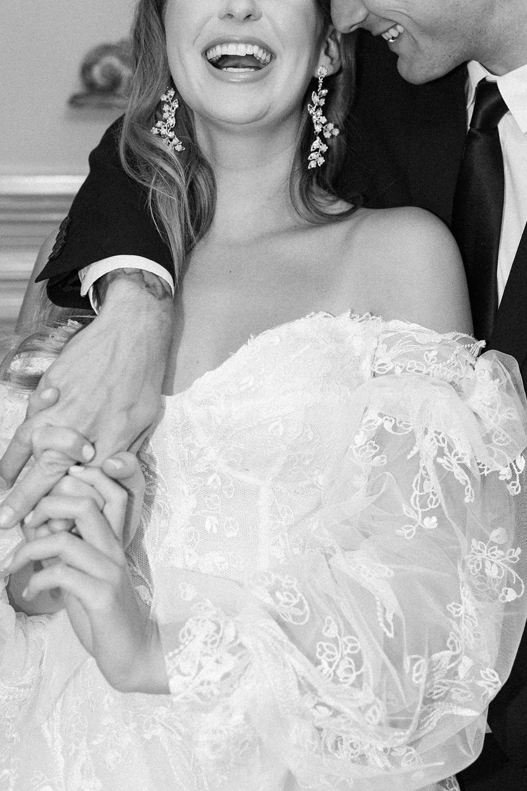 A joyful bride and groom holding hands, smiling, with the bride wearing a lace wedding gown and statement earrings, and the groom wearing a tuxedo.