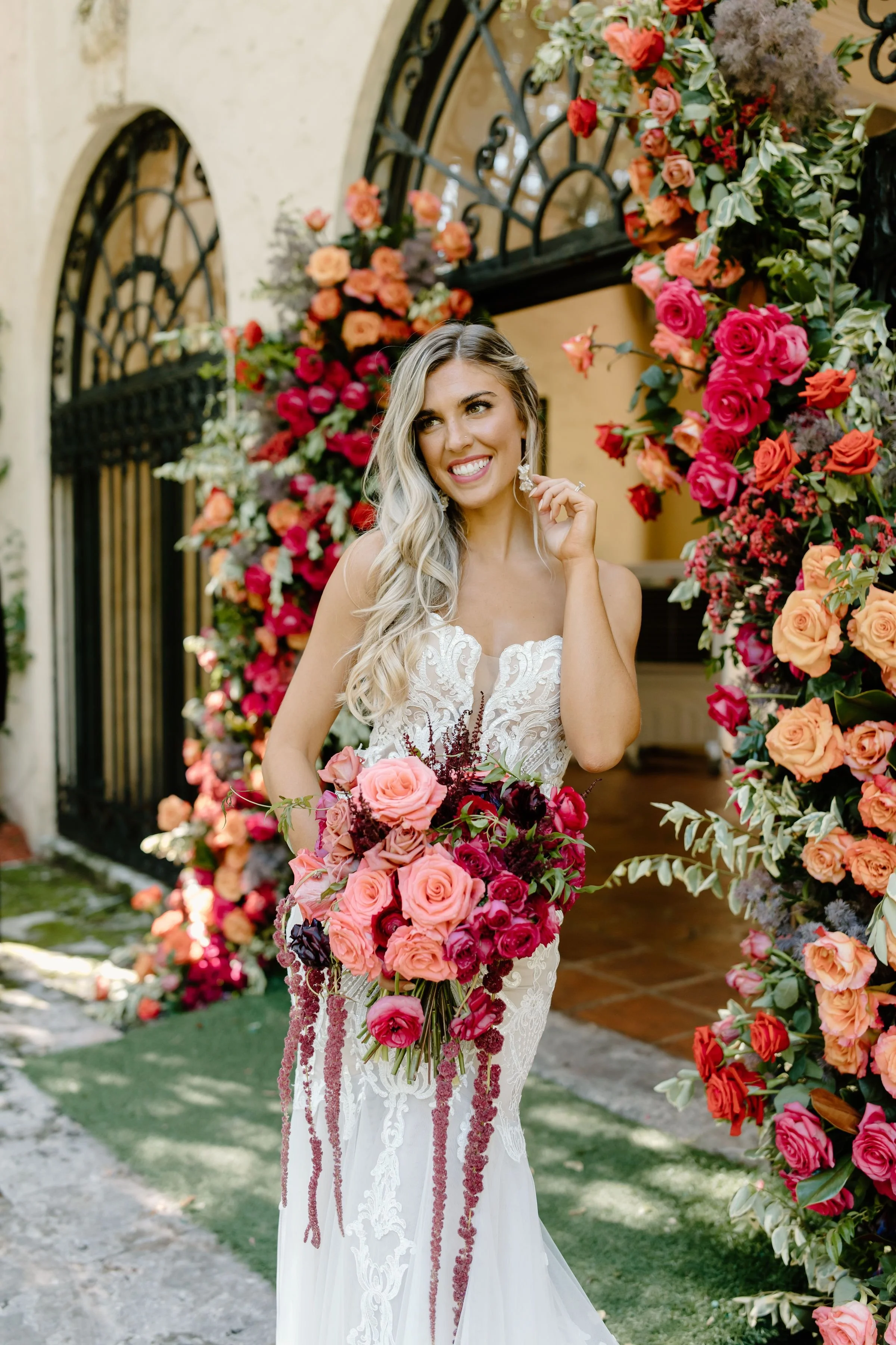 A bride in a white lace wedding gown holding a bouquet of pink and burgundy roses, standing in front of an archway decorated with pink, purple, and peach flowers.