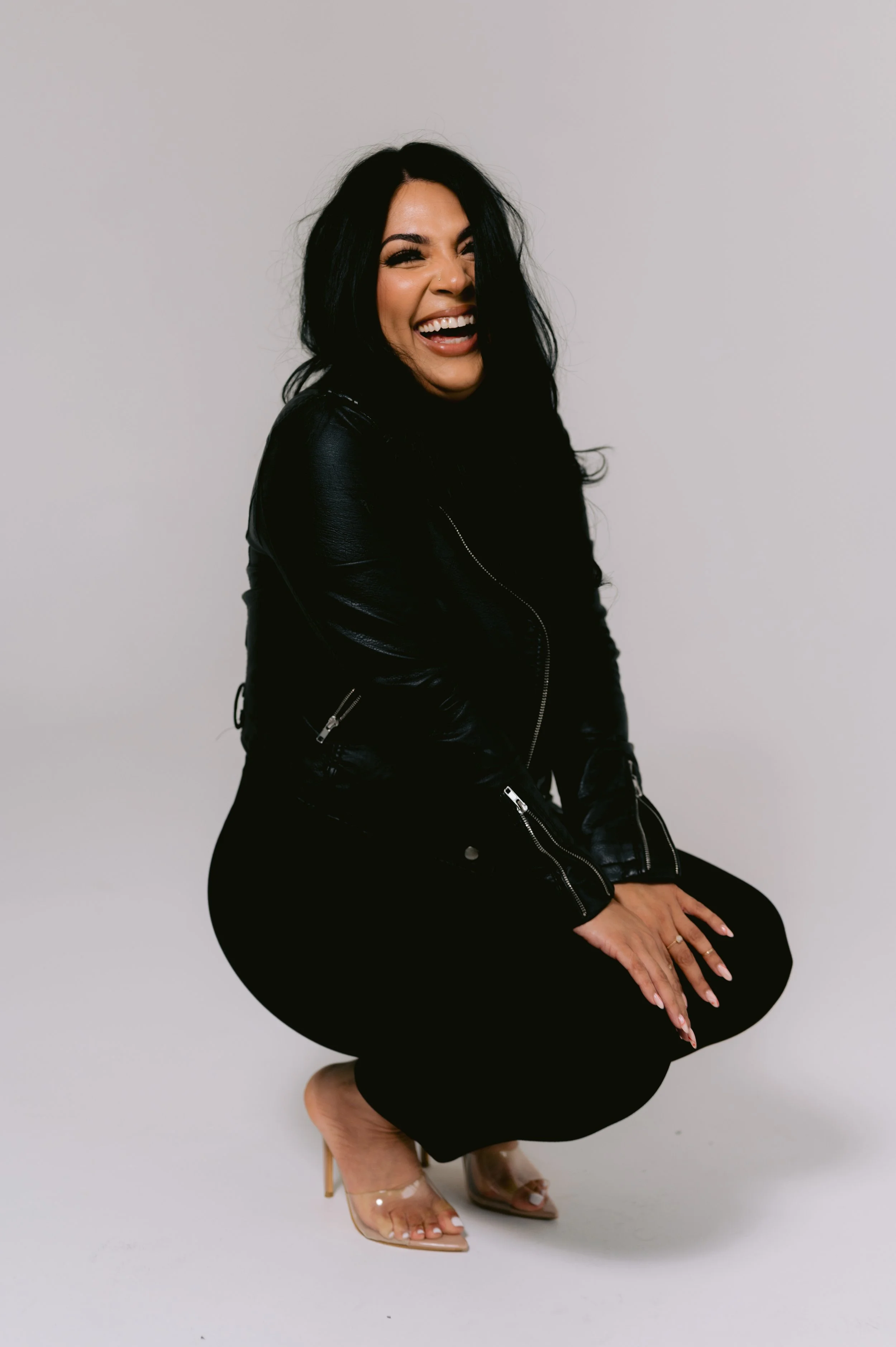 Woman in black leather jacket and black pants crouching, smiling and laughing, wearing clear high heels, with long black hair and light makeup, against a plain white background.