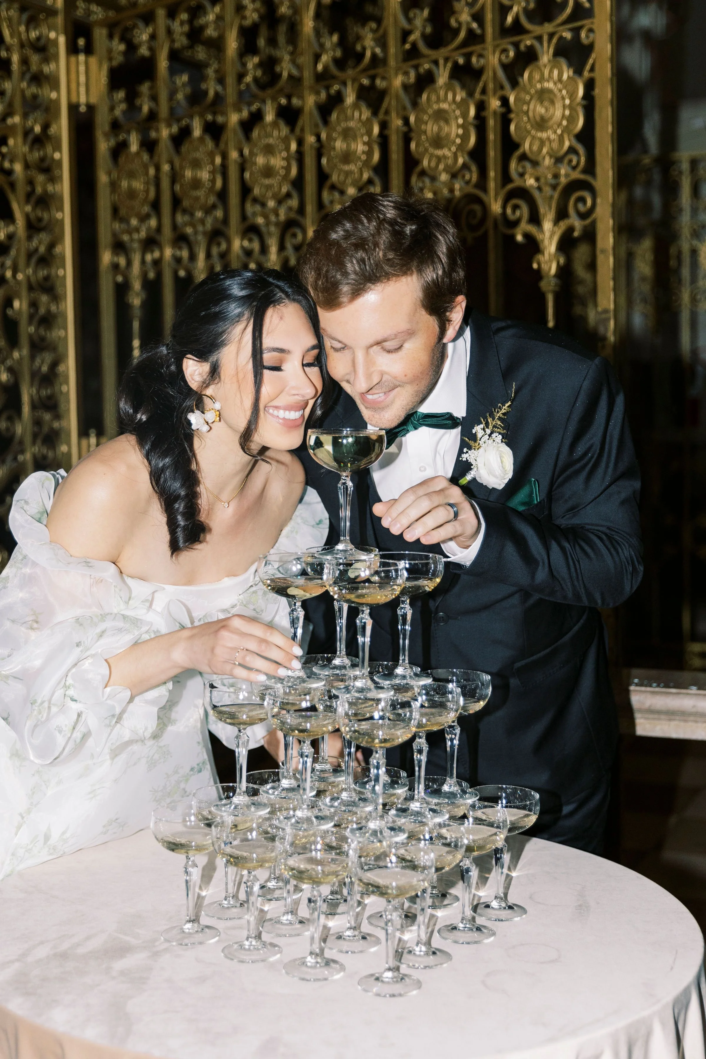 A bride and groom at their wedding reception, toasting with champagne glasses in a pyramid formation, smiling and dressed in wedding attire, with a decorative gold backdrop.