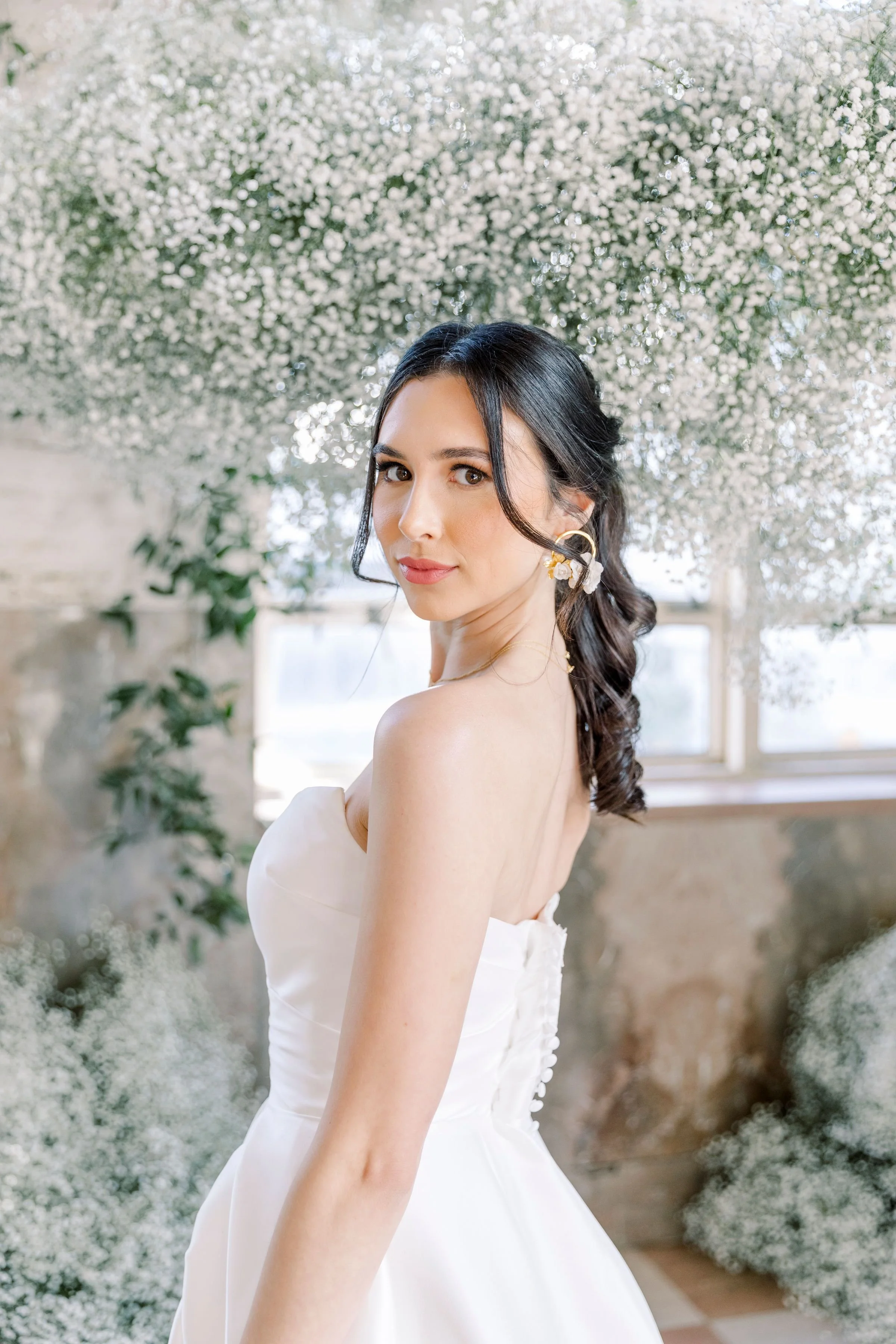 A bride with. soft wedding glam in a white strapless wedding dress standing in front of a background of white flowers and greenery.