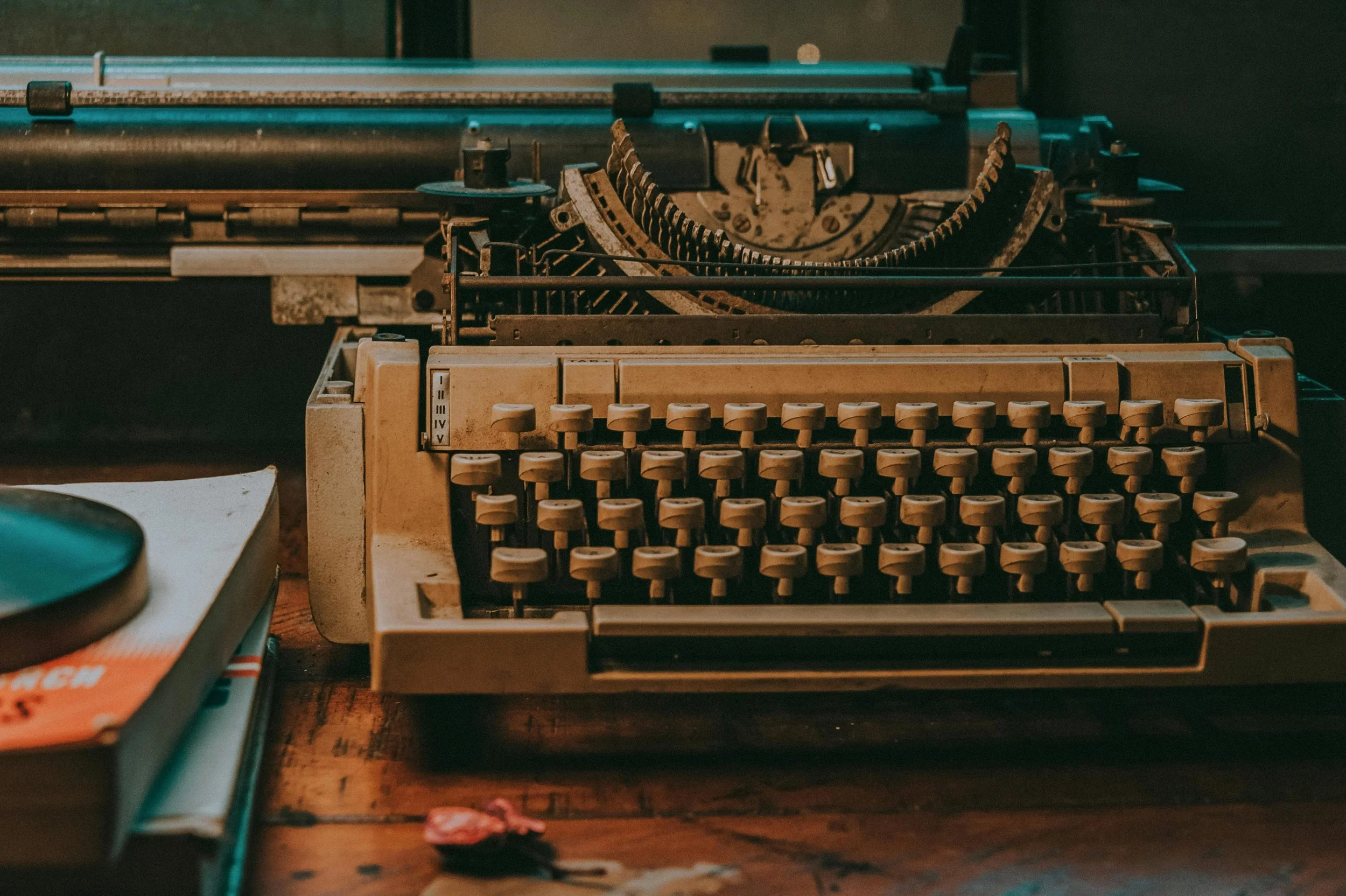 An old beige typewriter on a wooden desk, with some books and a small item nearby.