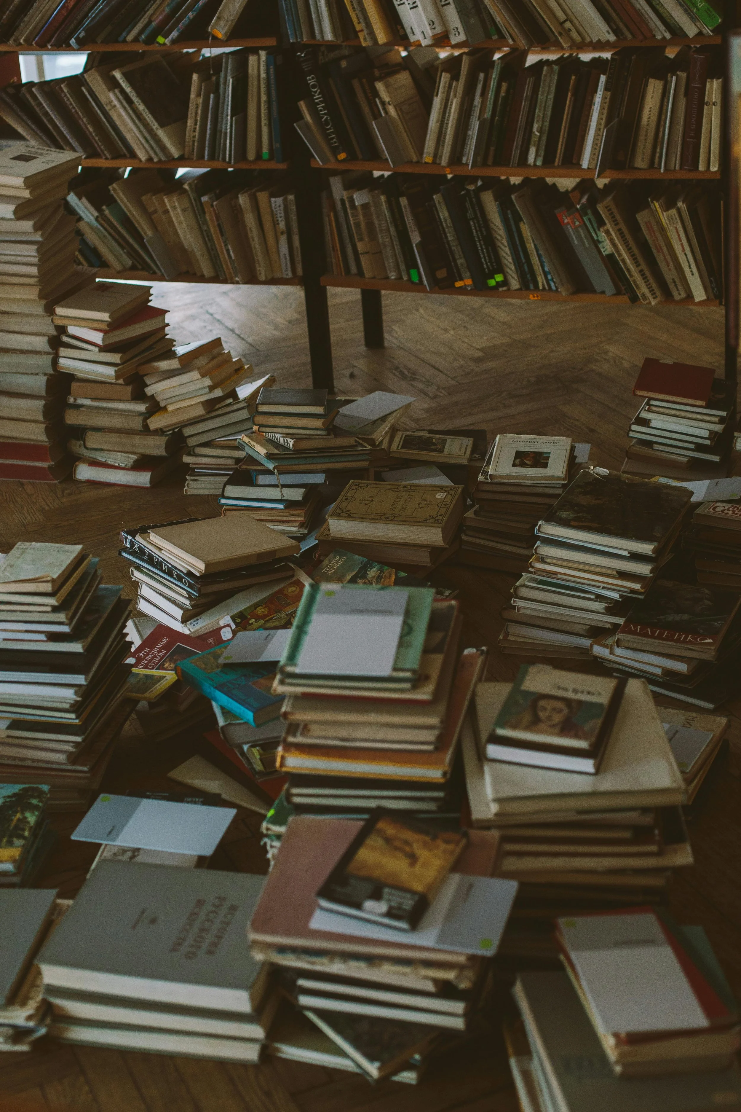 Many piles of books on the floor and a bookshelf filled with books in the background