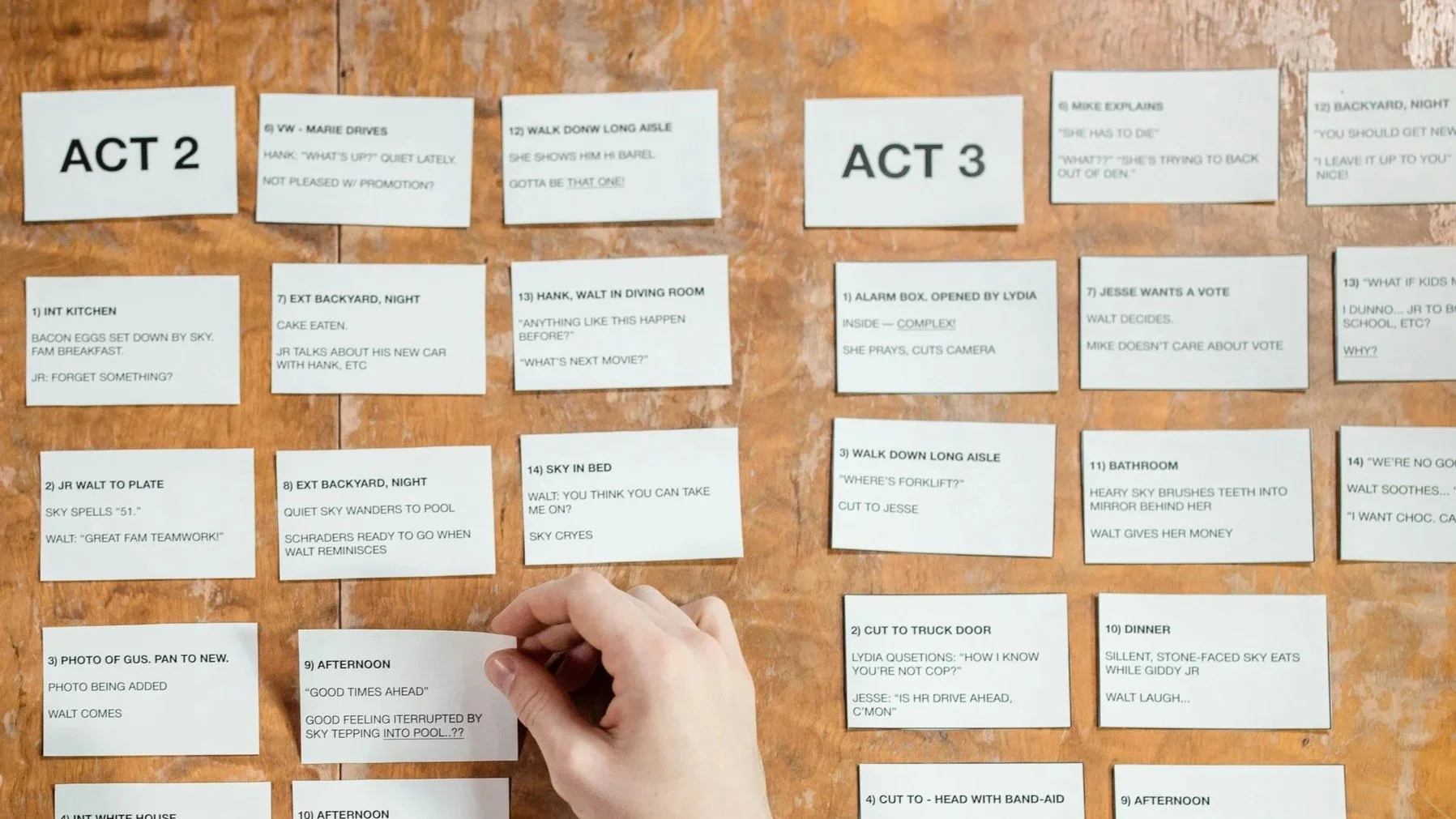 A person placing a white paper note on a wooden surface with several other handwritten notes arranged in a grid, labeled with acts and scenes.