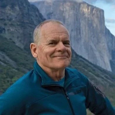 A man with short gray hair smiling outdoors in front of mountains and cliffs.