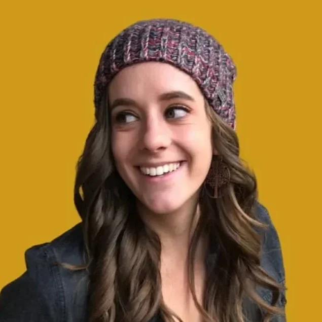 A young woman with long, wavy brown hair wearing a colorful knit beanie and a dark denim jacket, smiling and looking to her right against a yellow background.