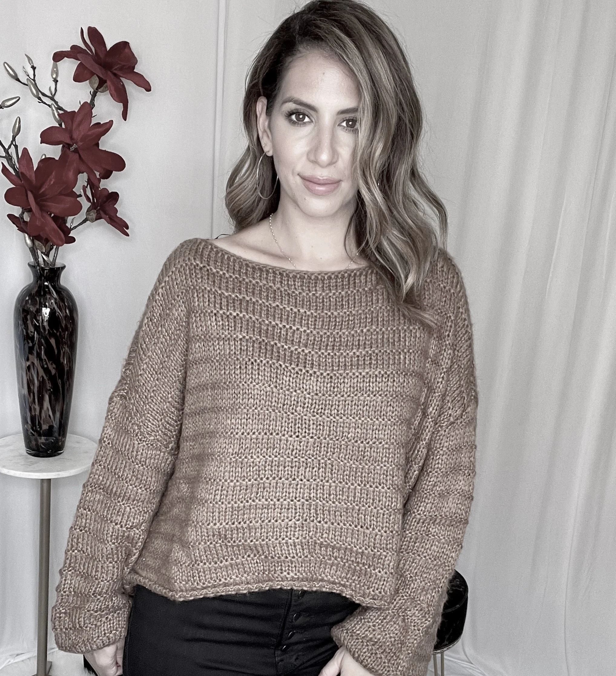A woman with shoulder-length wavy hair and hoop earrings standing indoors near a side table with a black vase containing red flowers, wearing a tan knitted sweater and black pants.