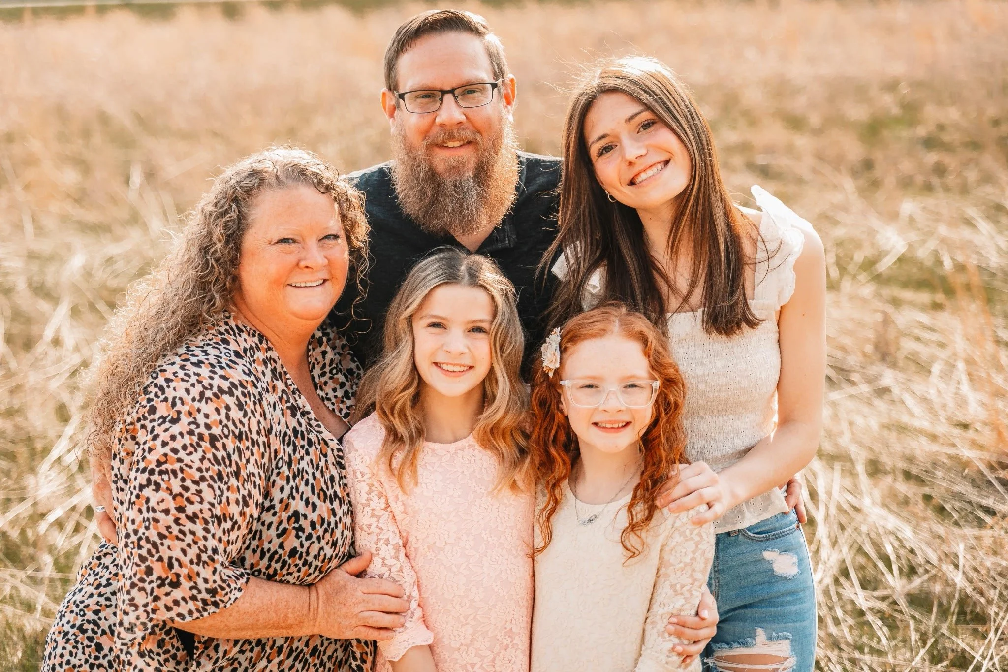 Family of six outdoors in a grassy field, smiling for a group photo. Includes an older woman, a man with glasses and a beard, a teenage girl, two young girls with red hair, one with glasses, and a woman with long brown hair.