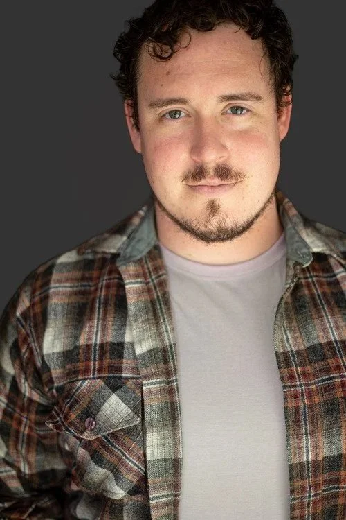 A young man with curly hair and a light beard, wearing a plaid shirt over a white T-shirt, standing against a dark background.