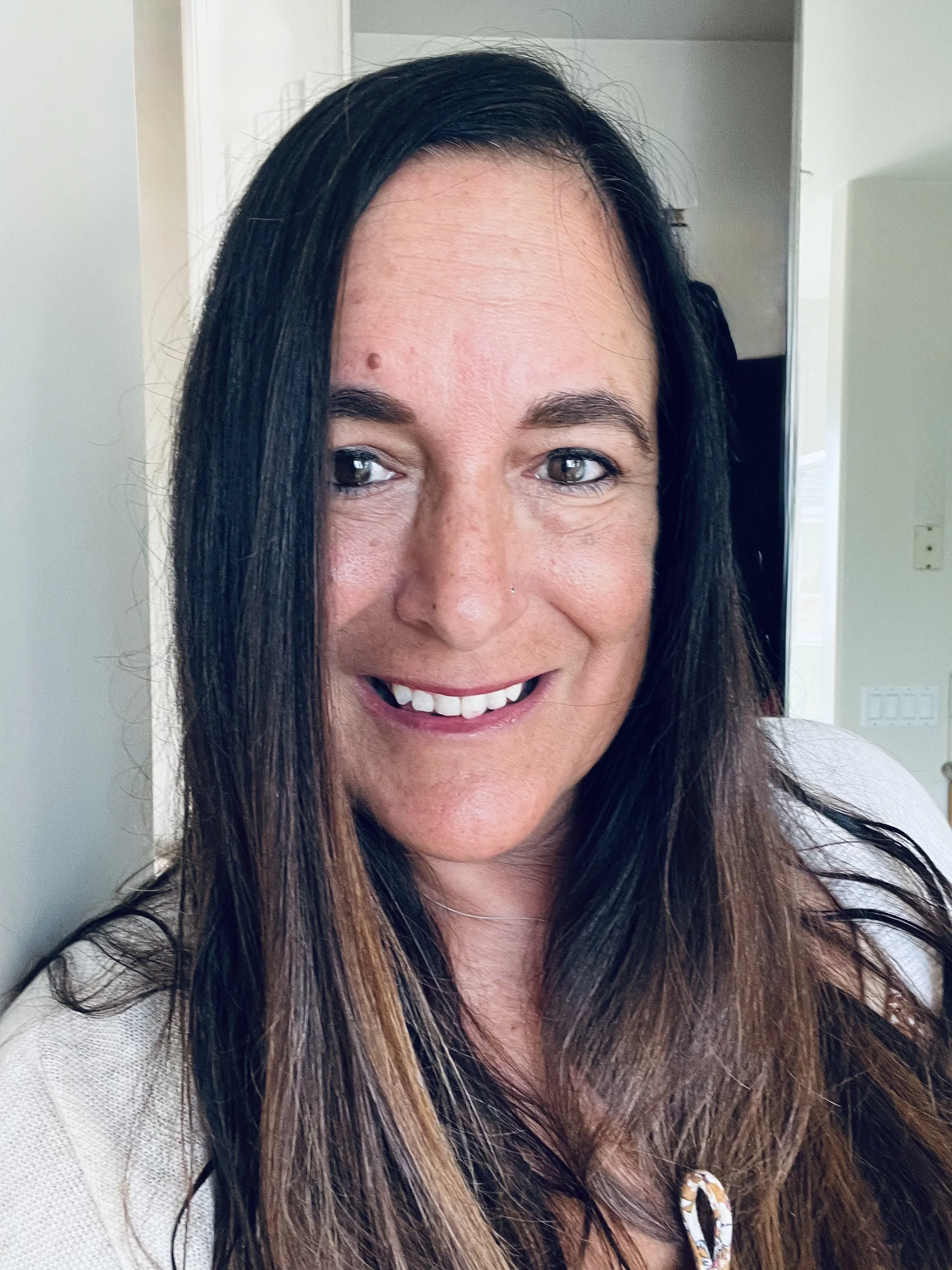 A smiling woman with long dark hair, wearing a white top, standing indoors with a neutral background.