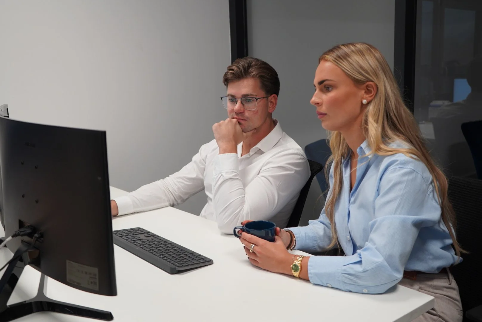 Twee mensen werken samen op een kantoor, een man met bril en een vrouw met lang blond haar, ze kijken aandachtig naar een computerscherm. De vrouw legt de man persoonlijk financiele zaken uit.