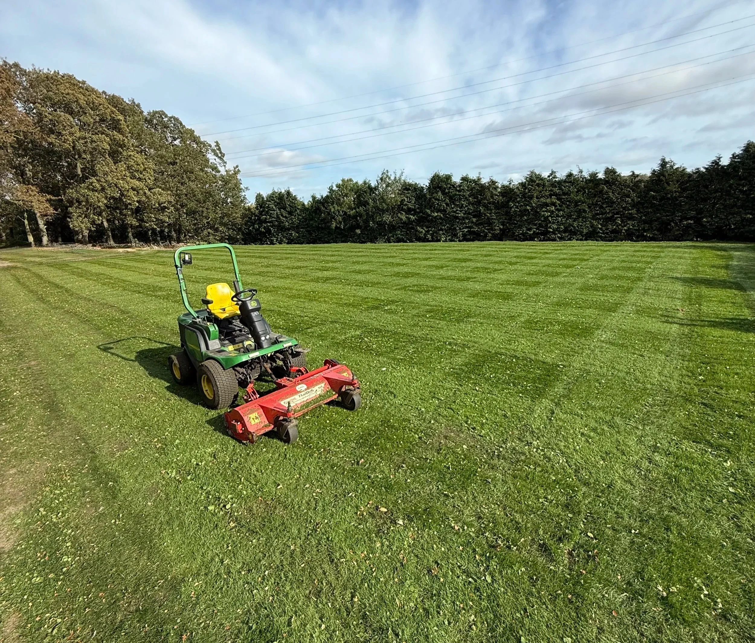 A grass cutting machine in an open grassy field with trees in the background and a partly cloudy sky.