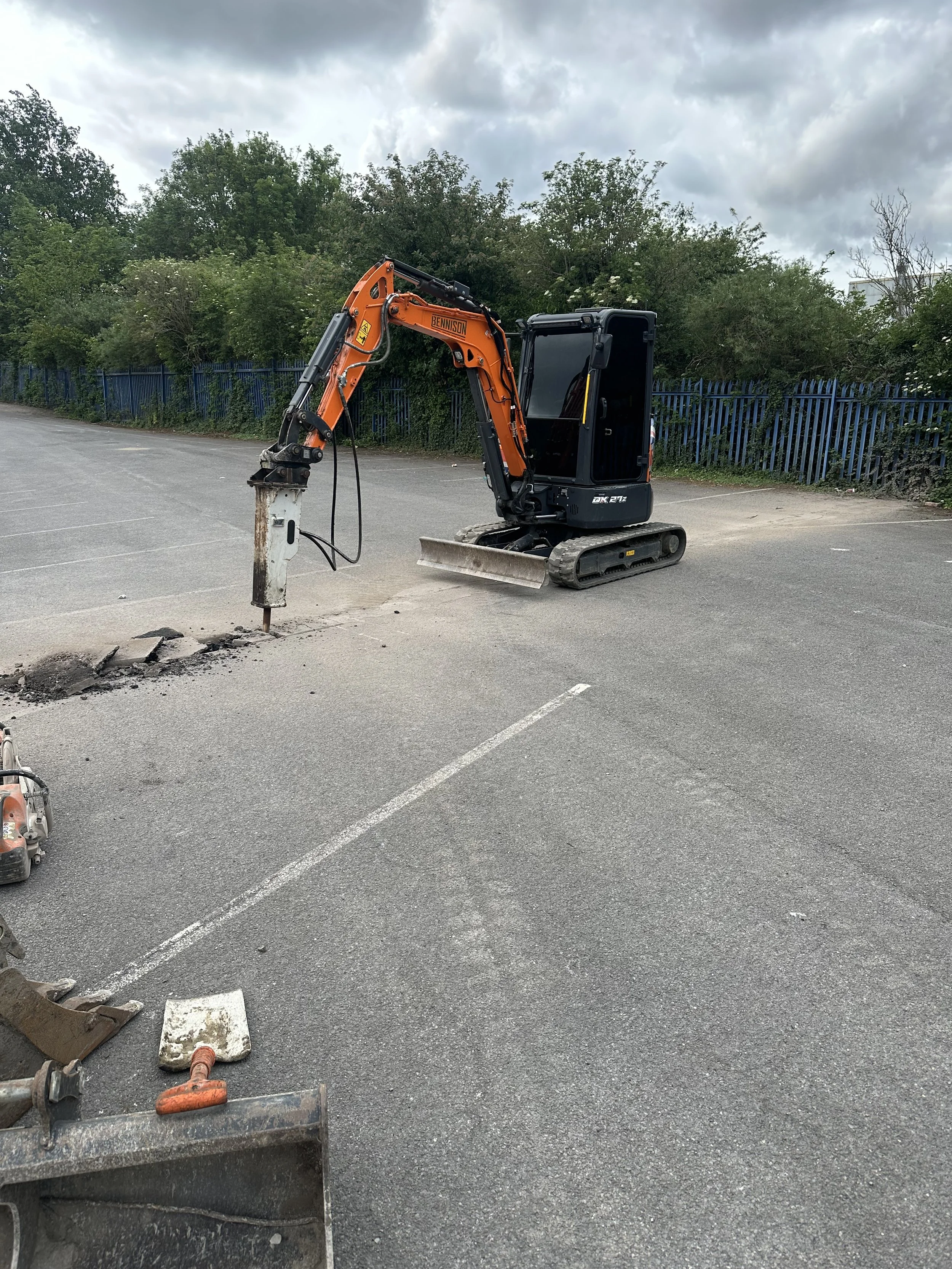 A small orange and black excavator breaking up a section of asphalt parking lot. The sky is cloudy, and there is a blue fence with trees behind it.