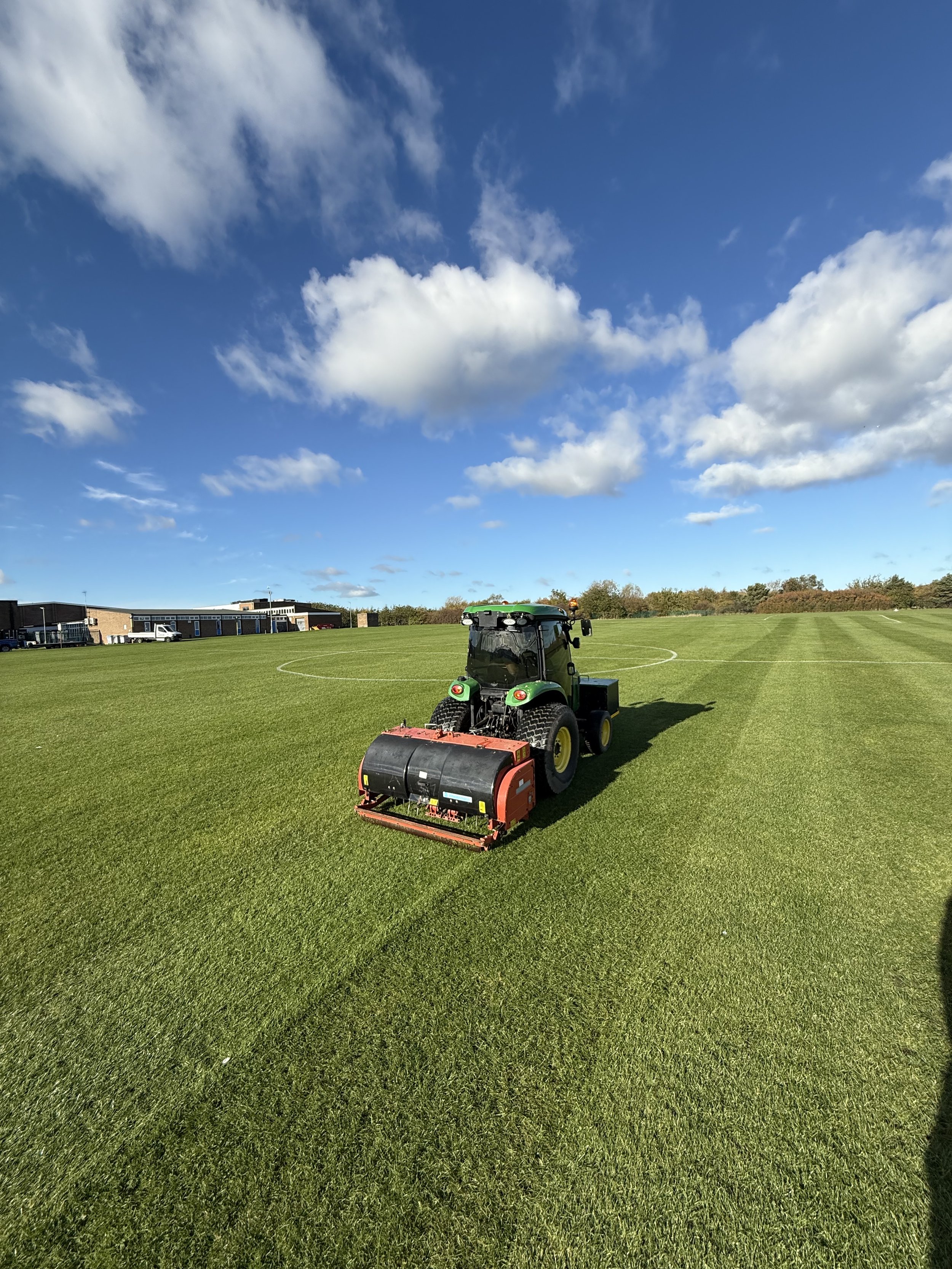 A green tractor with a large roller attachment working on a well-maintained grass field under a partly cloudy blue sky.