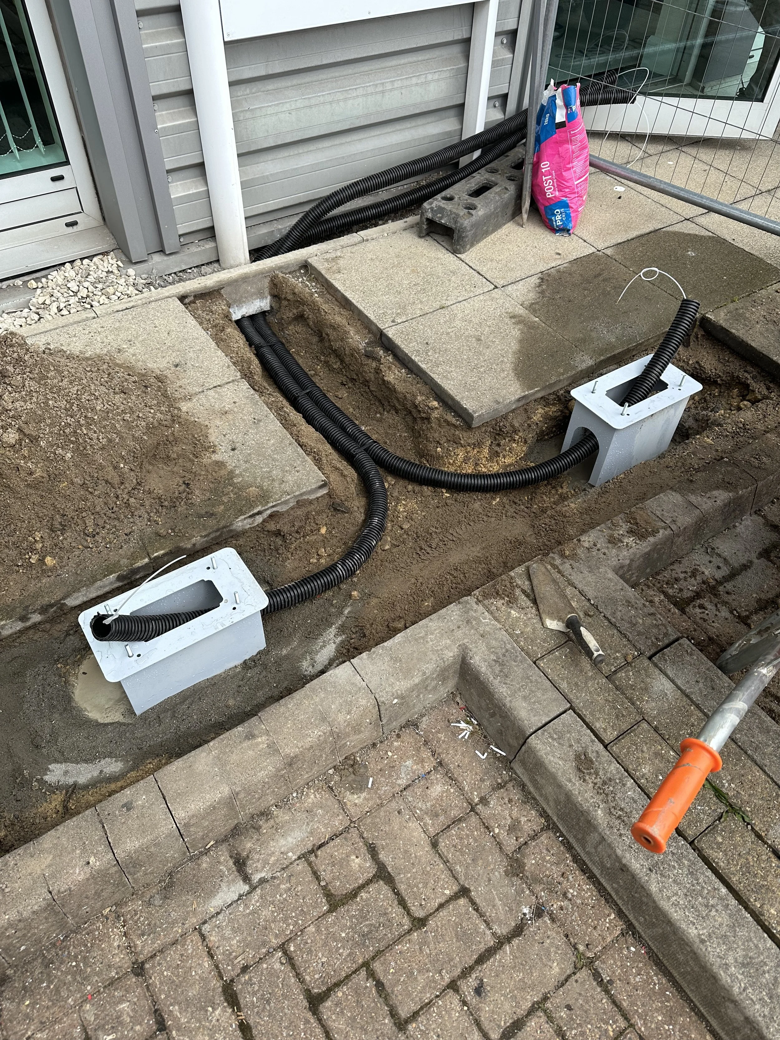 Construction site with buried conduits and cables for an electrical or communication system installation, with concrete blocks, bricks, and tools visible.