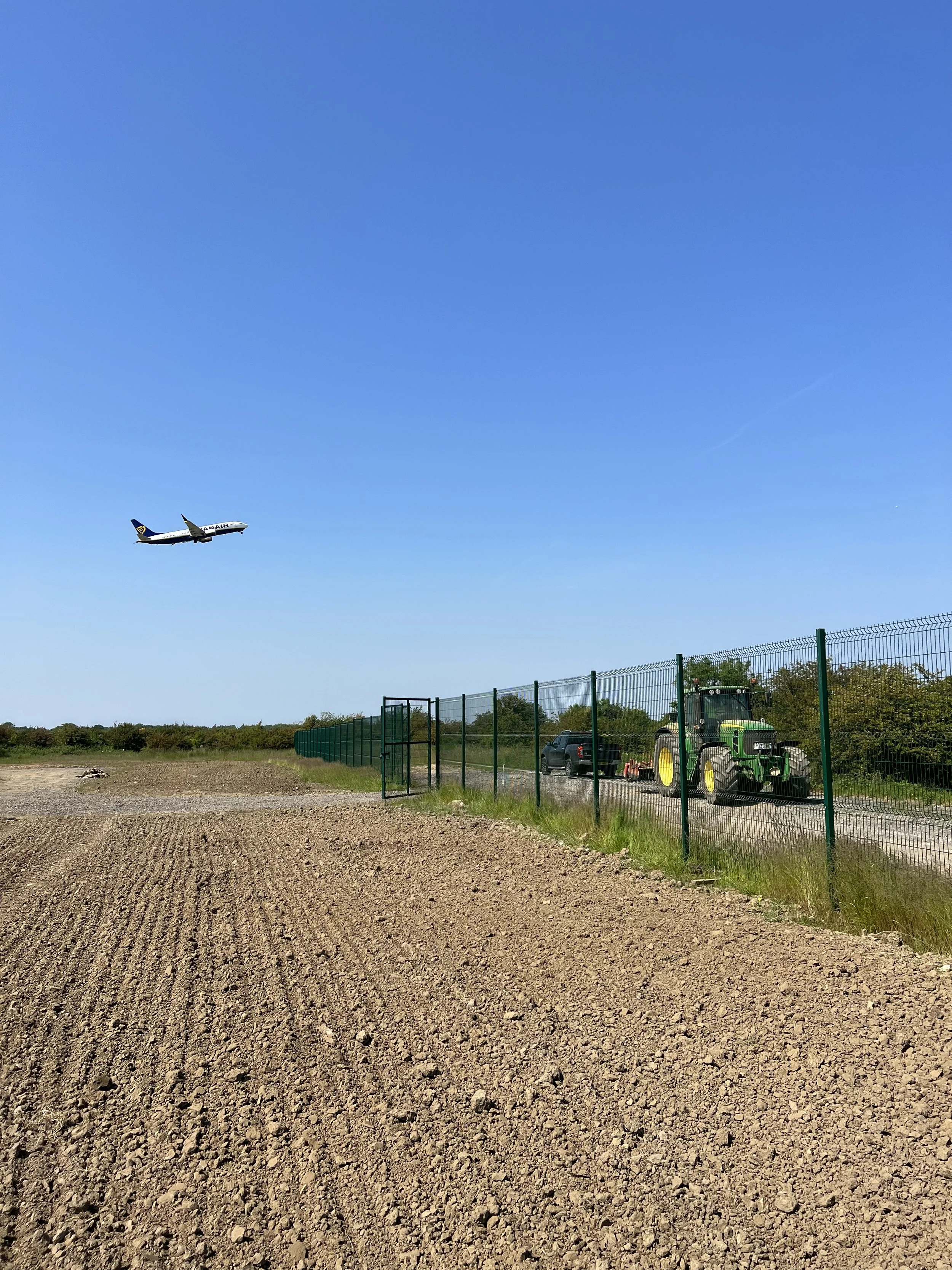 An airplane flying in the clear blue sky near a rural farmland with a tractor and truck parked by a green fence.