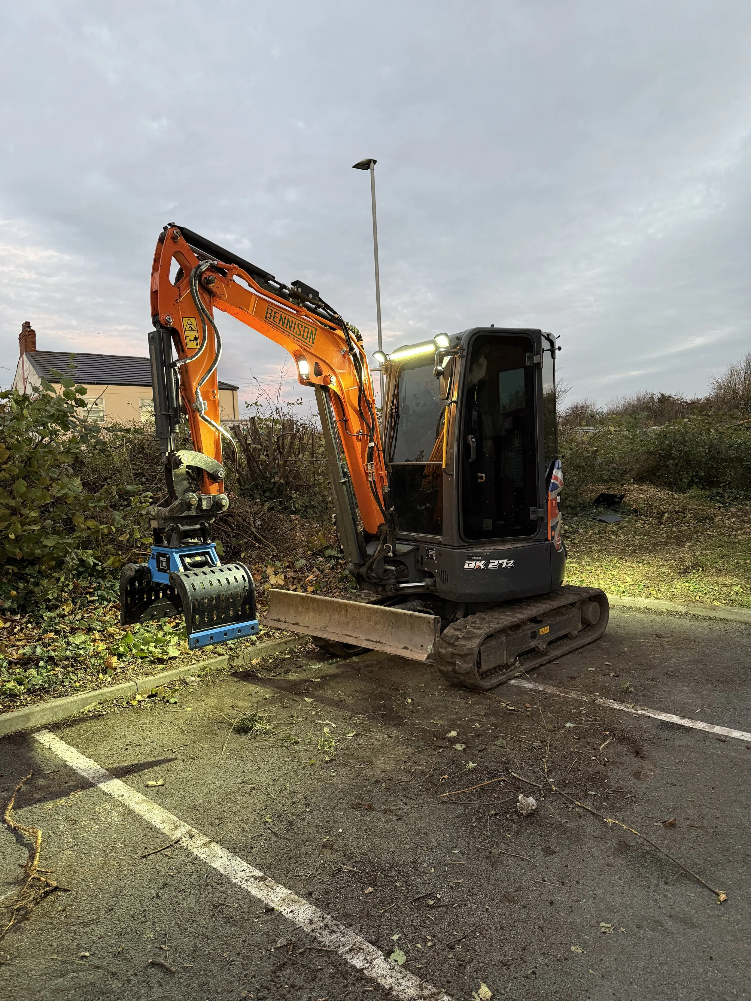 Mini excavator parked in a parking lot with overgrown bushes nearby, working on clearing vegetation