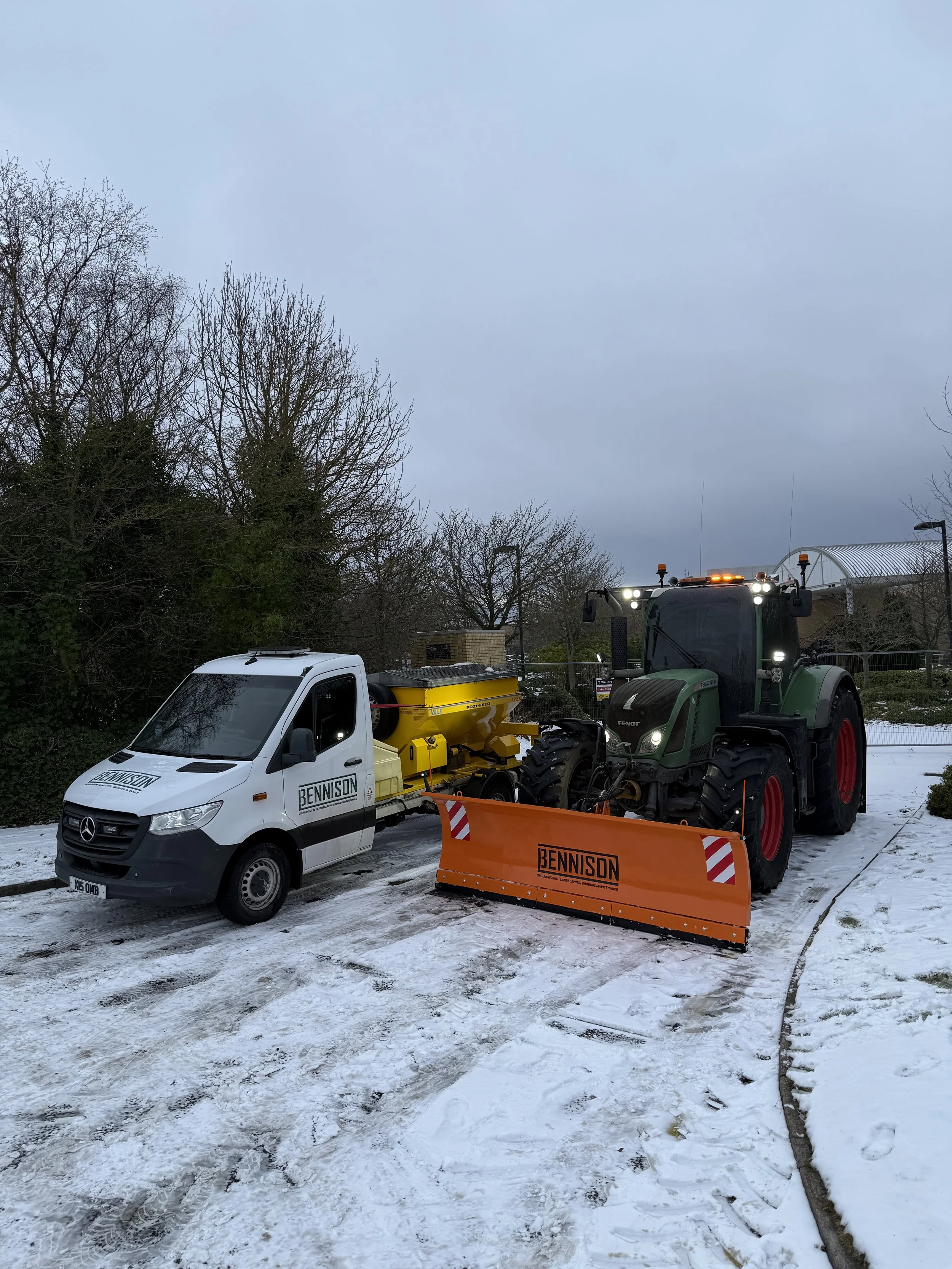 A tractor and a van, both marked with 'BENNISON', parked on a snow-covered path with leafless trees in the background.