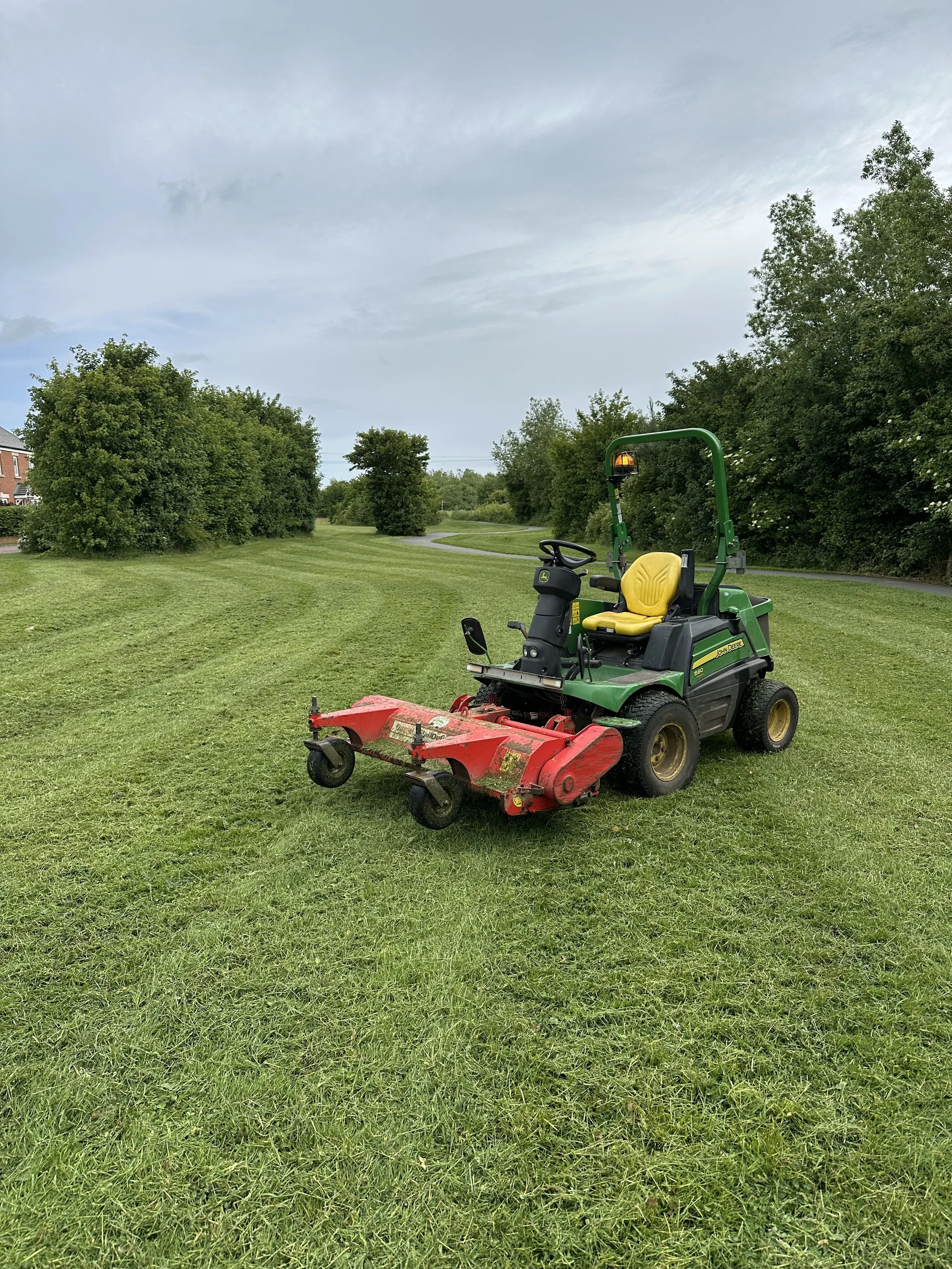 Green and black lawn mower with yellow seat on a grassy field with trees in the background and cloudy sky.