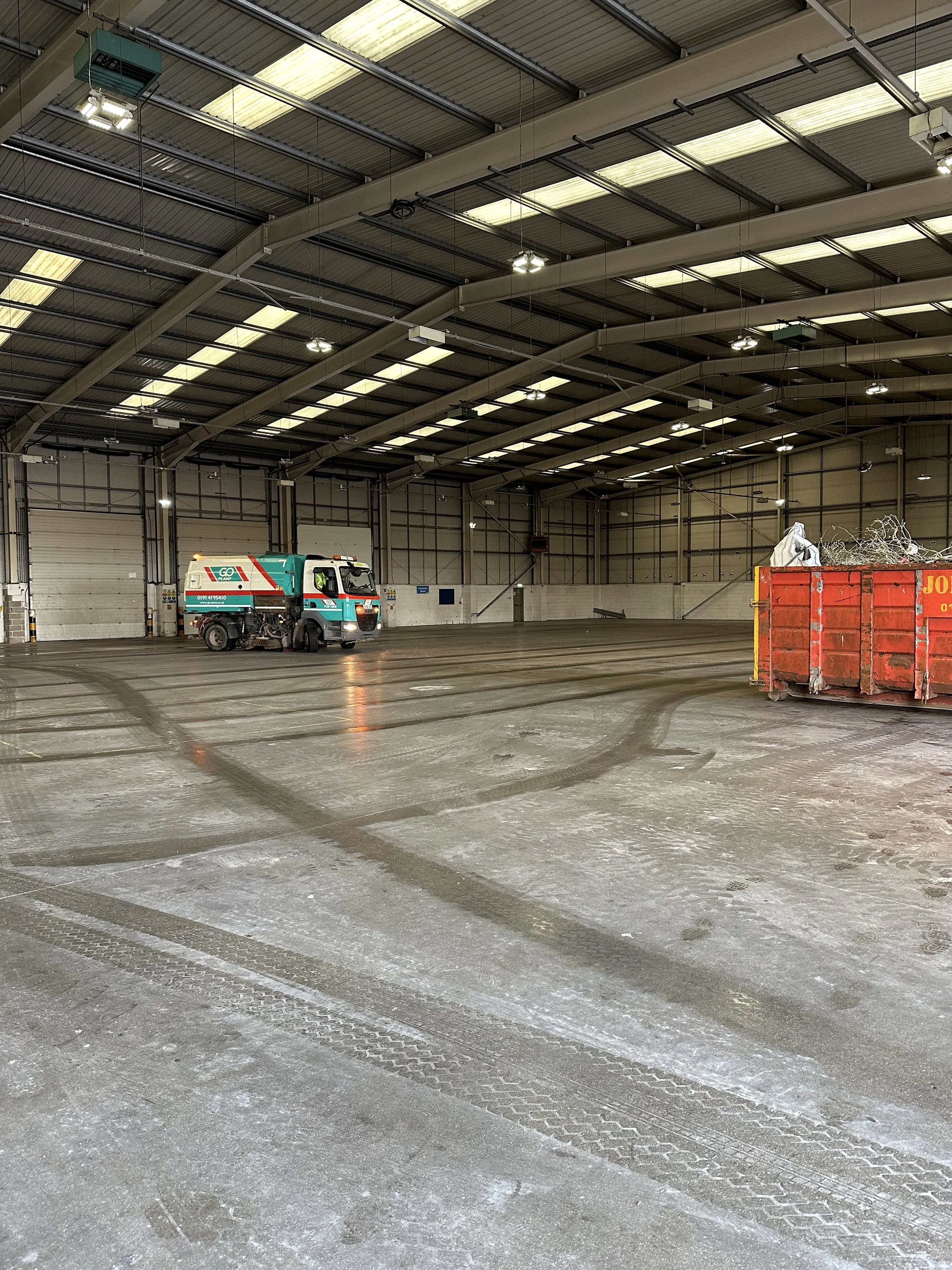 Empty warehouse with a floor cleaning machine and a red dumpster filled with construction debris.
