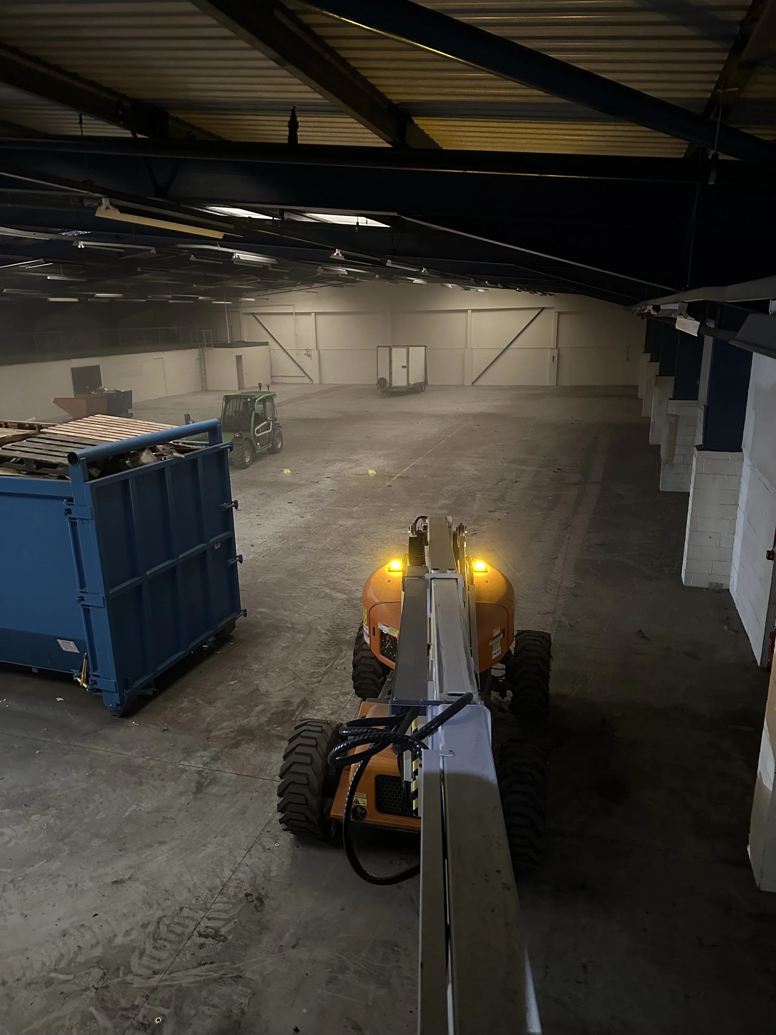 Inside a large warehouse with high ceilings, a small orange construction vehicle with a long arm is positioned front and center. To the left, there is a blue dumpster and a small green utility vehicle. In the background, there are empty open spaces and a small cart, with some yellow markers on the floor.