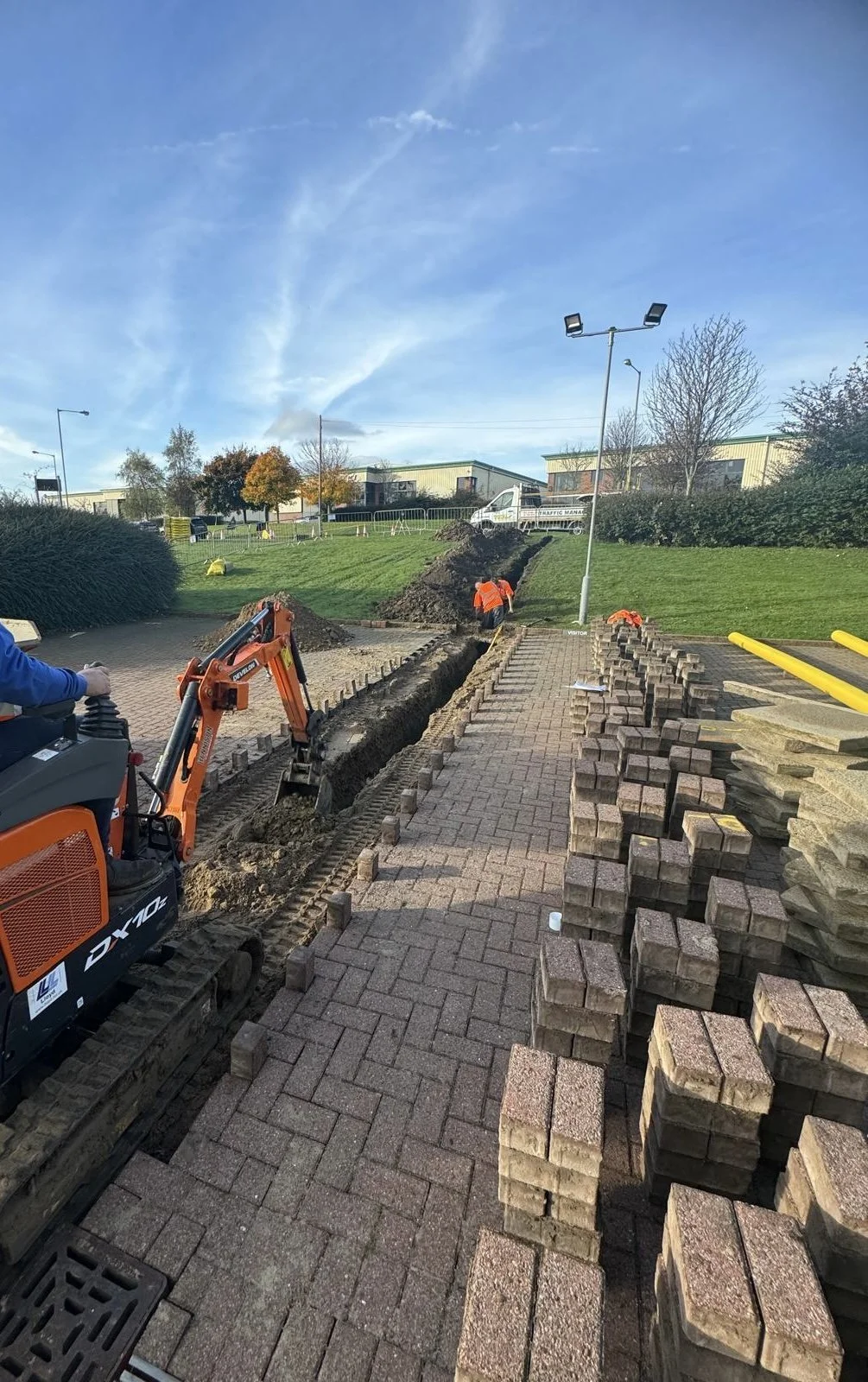 Workers laying bricks and installing bricks on pathway as part of construction or renovation project outdoors, with stacks of bricks on the side.