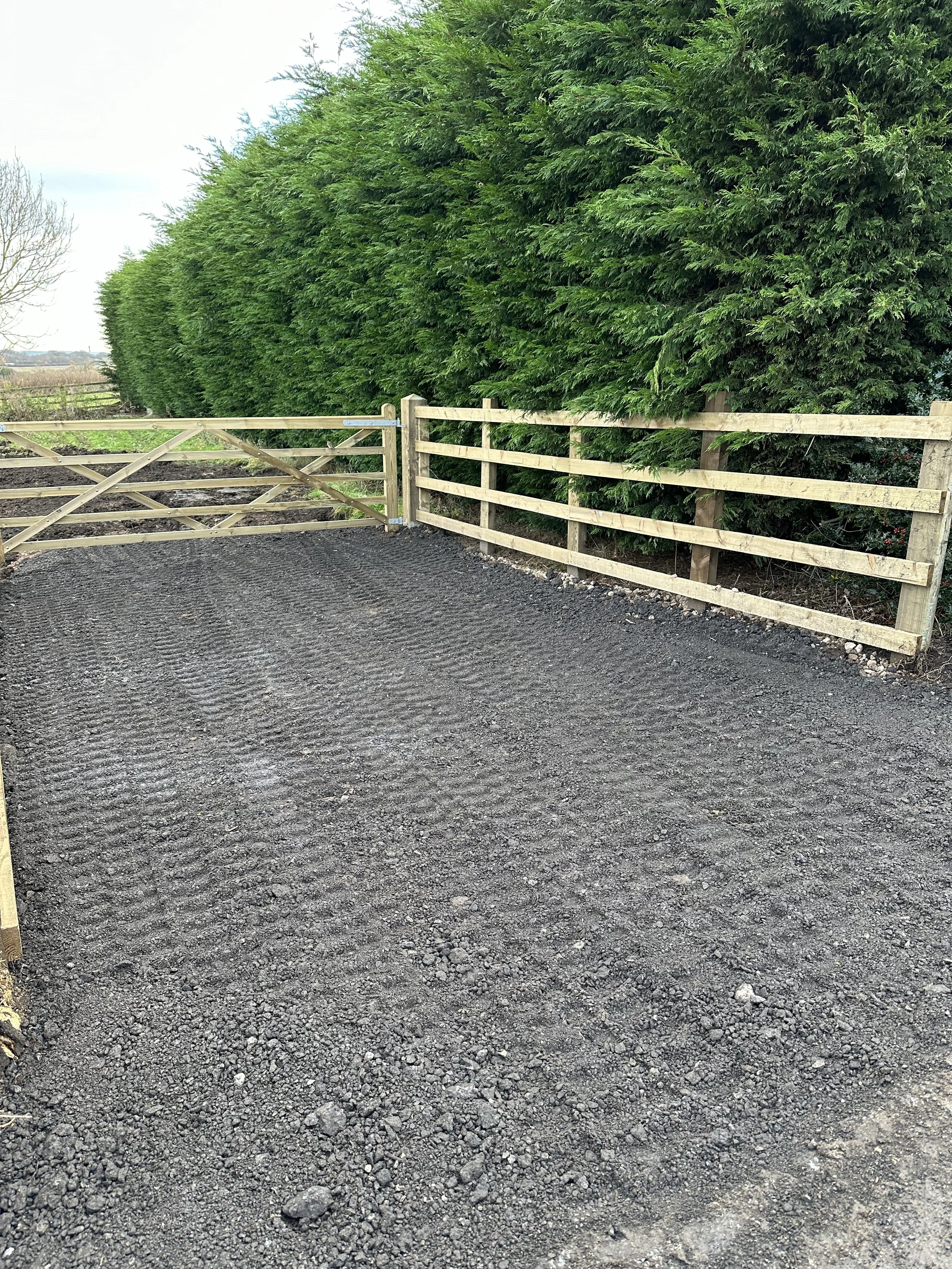 A gravel pathway bordered by a wooden fence on one side and a dense line of green trees on the other, with an open gate in the fence.
