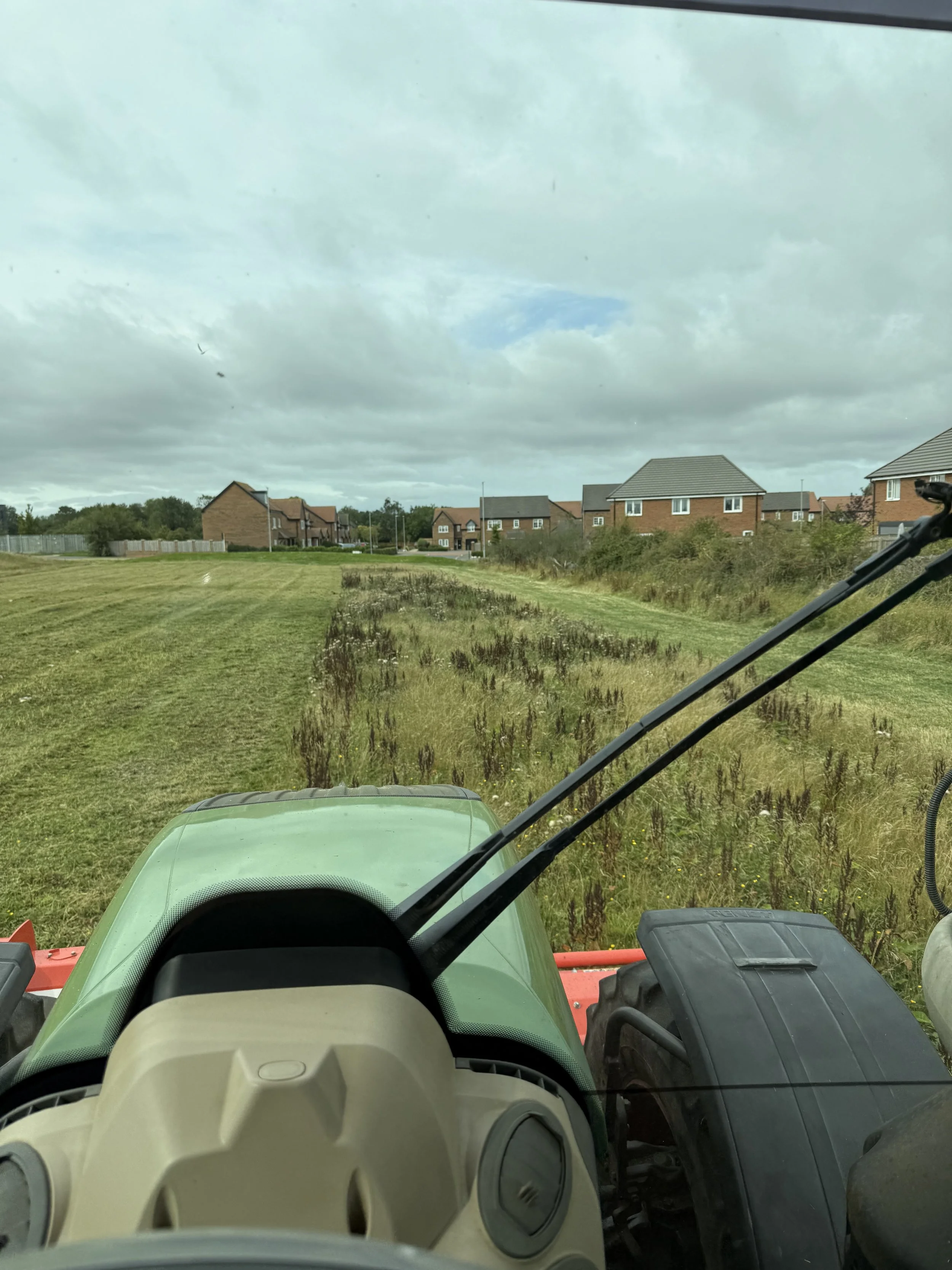 View through the windshield of a green tractor showing a grassy field with patches of taller grass and houses in the distance under a cloudy sky.