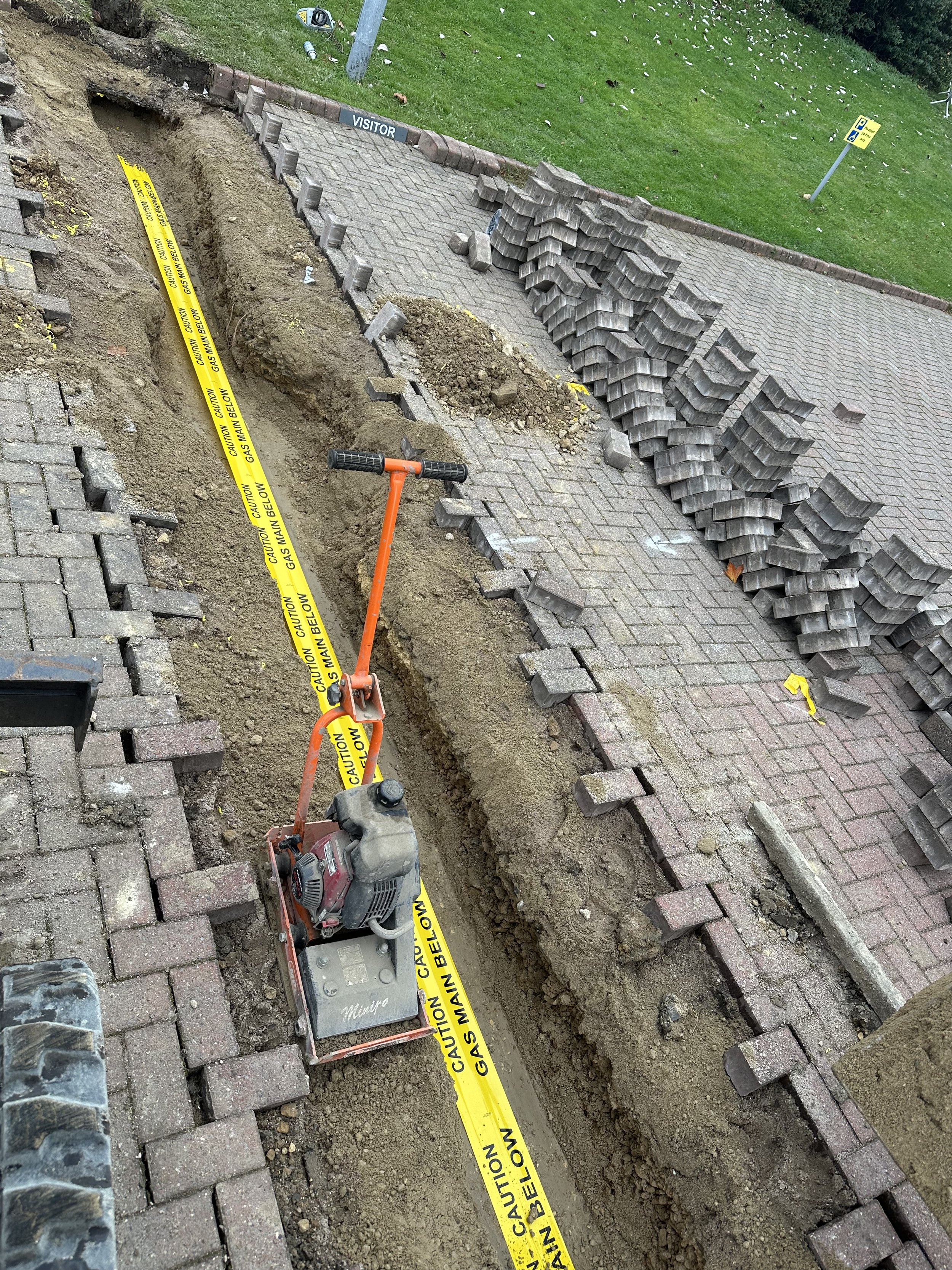 Construction site with a trench dug in a brick-paved sidewalk, caution tape warning about gas line, and a small compactor machine for soil, with stacked bricks nearby.