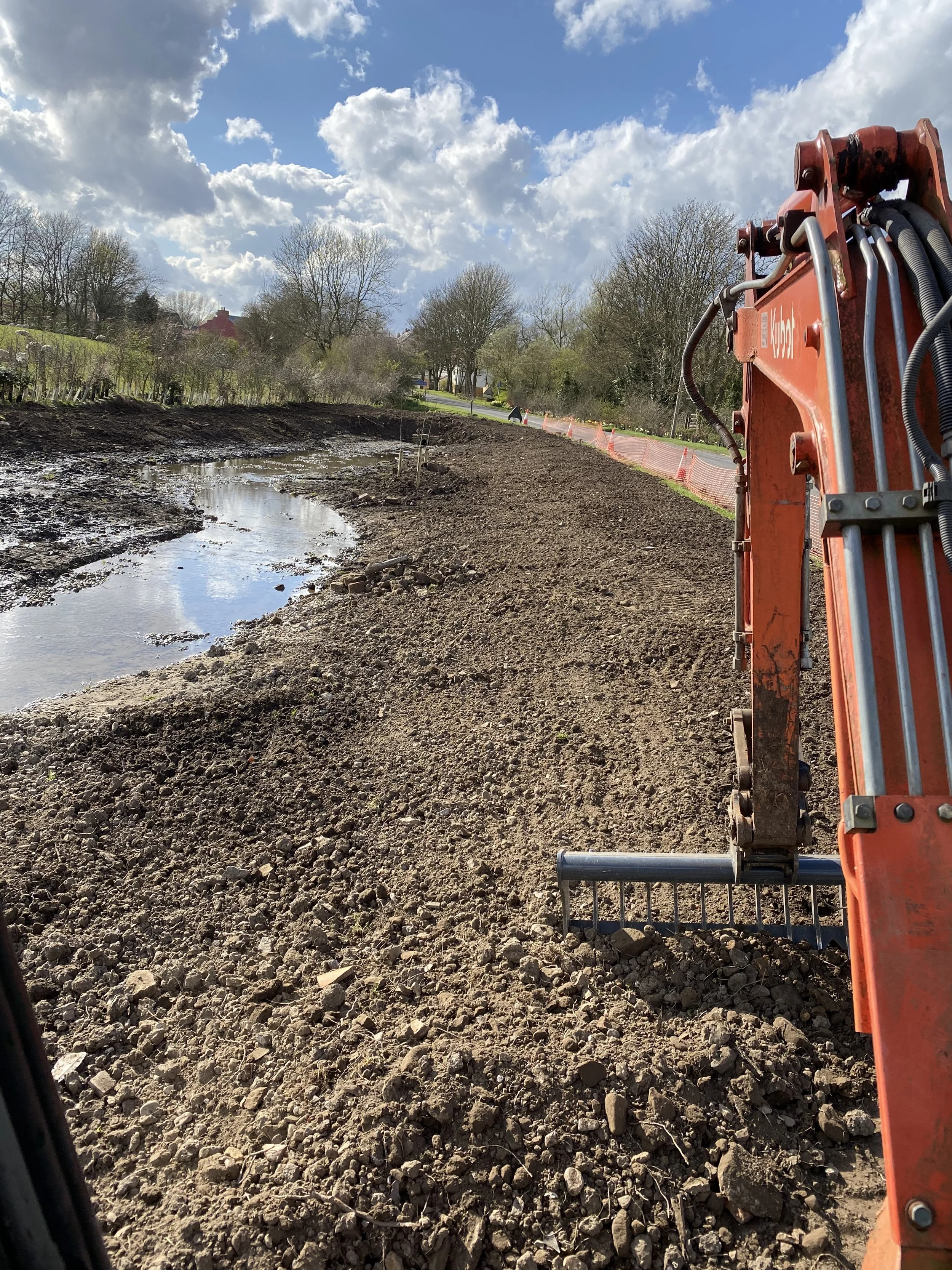 Construction site with a large orange excavator along a dirt pathway, with a small muddy pond on the left, under a partly cloudy sky.