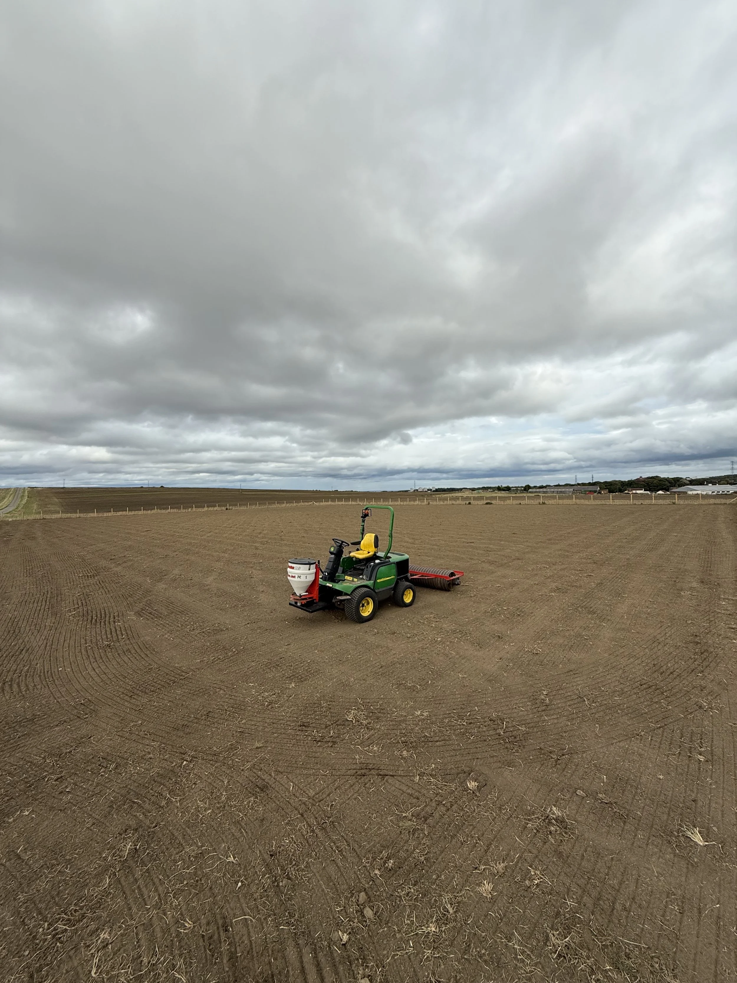 A small green and yellow tractor with a red attachment sits on a large, freshly tilled field under a cloudy sky.
